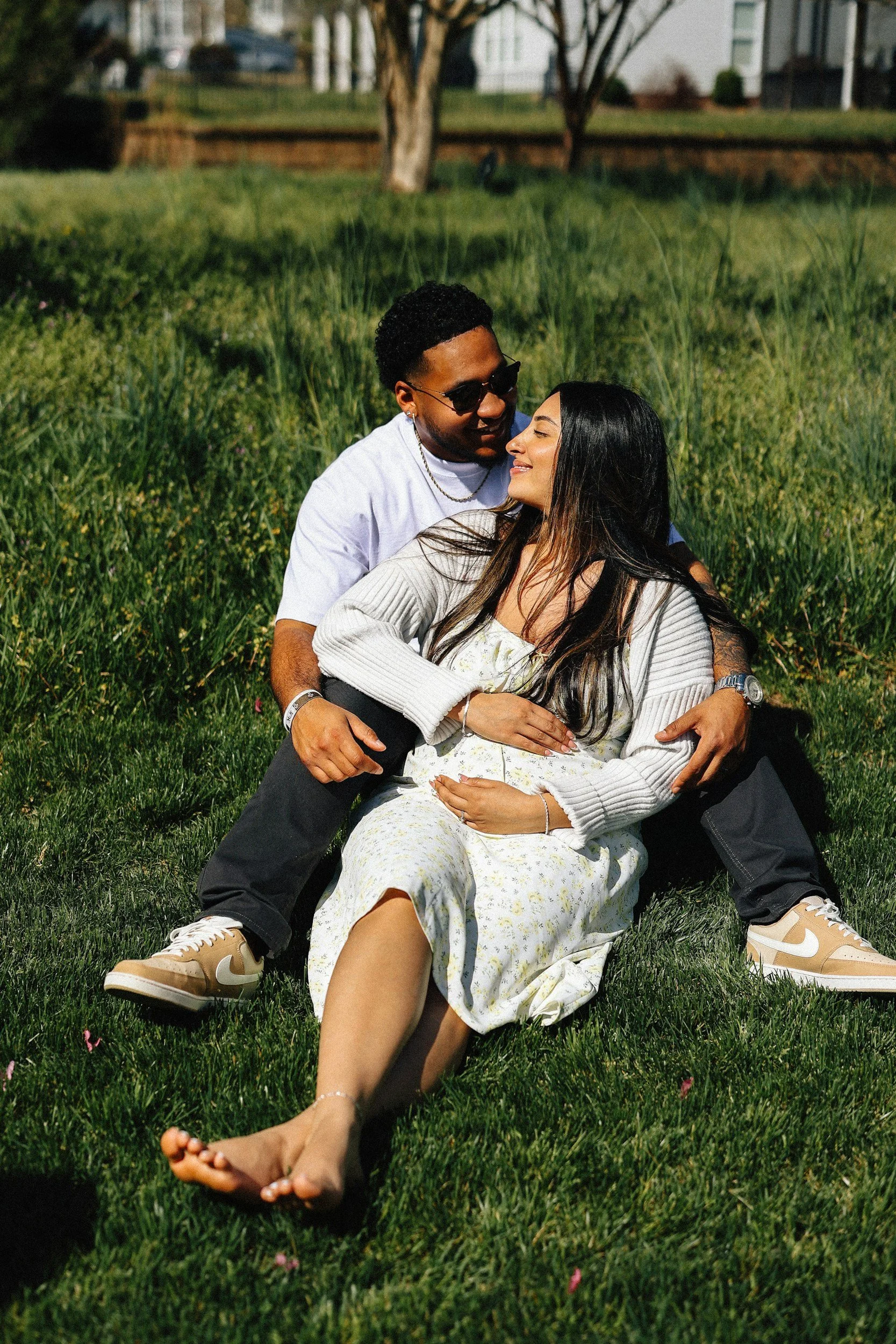 A young man and woman sitting on grass in a park, smiling and embracing each other. The man wears sunglasses, a white t-shirt, and tan sneakers, while the woman wears a white dress with a light cardigan. The background includes trees and houses.