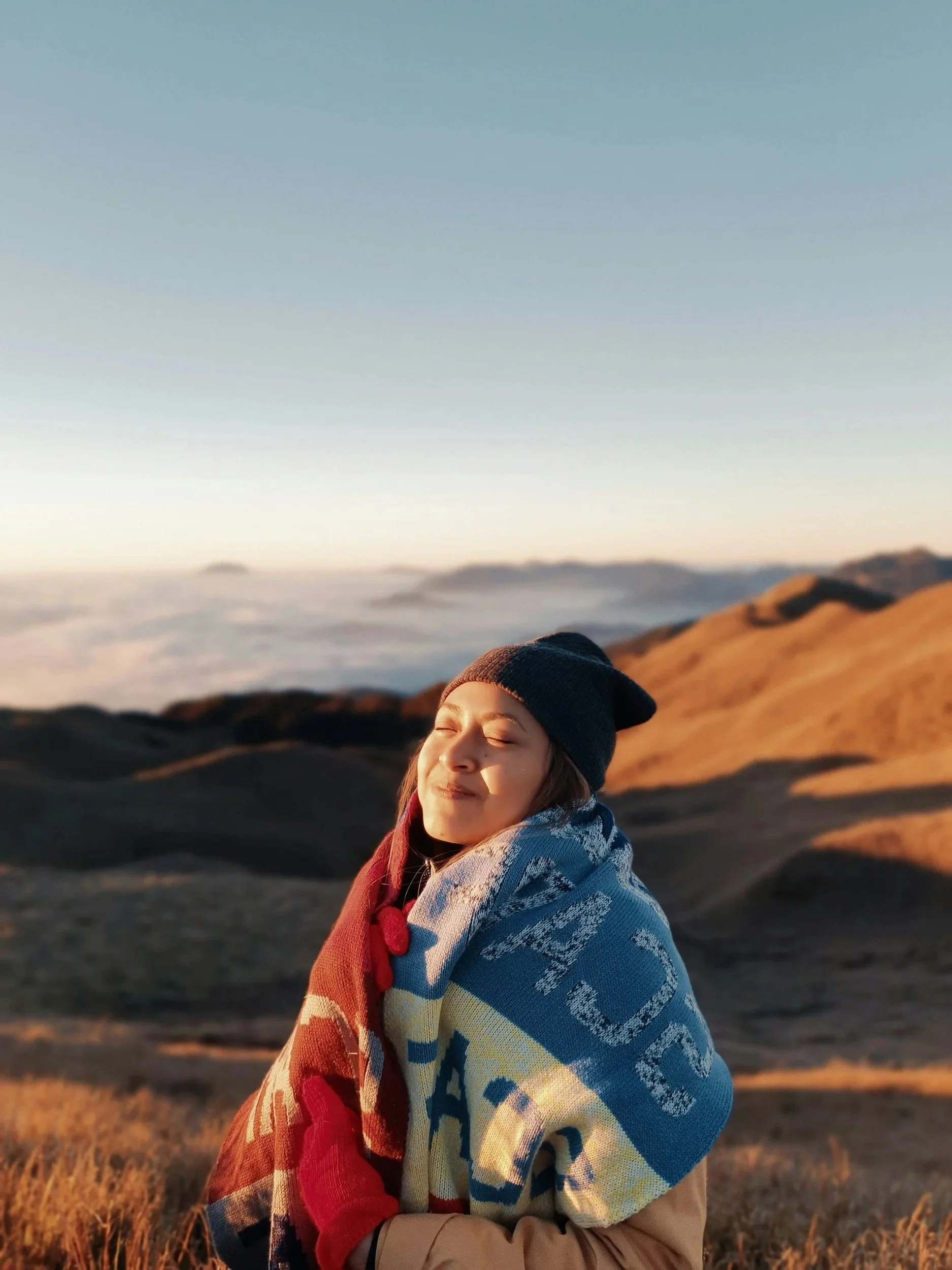 A person with closed eyes smiling, wrapped in a colorful blanket, standing in a field with mountains in the background during sunset.