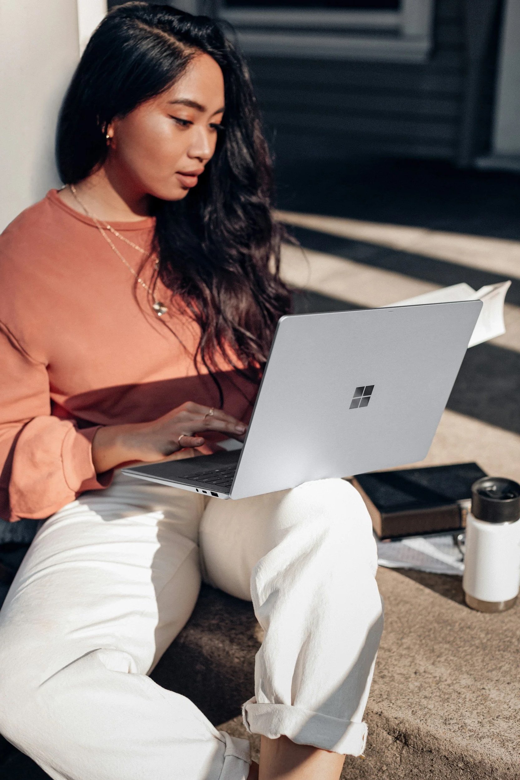 A woman sitting on the ground working on a silver Microsoft Surface laptop. She is wearing a peach-colored shirt, white pants, and gold jewelry, with long wavy black hair.