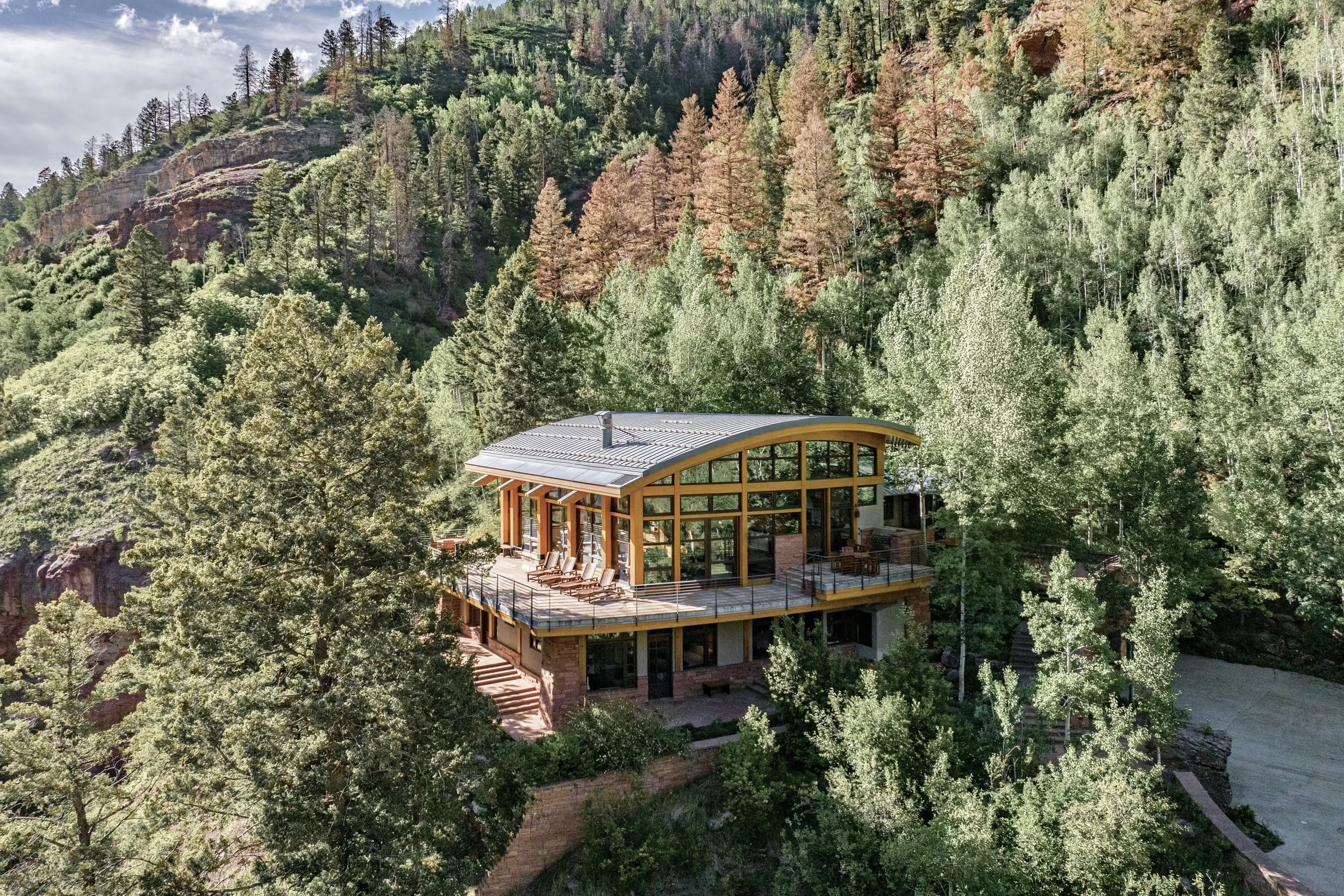 A modern house with large glass windows and a curved roof, built on a hillside surrounded by dense green forest and mountain terrain.