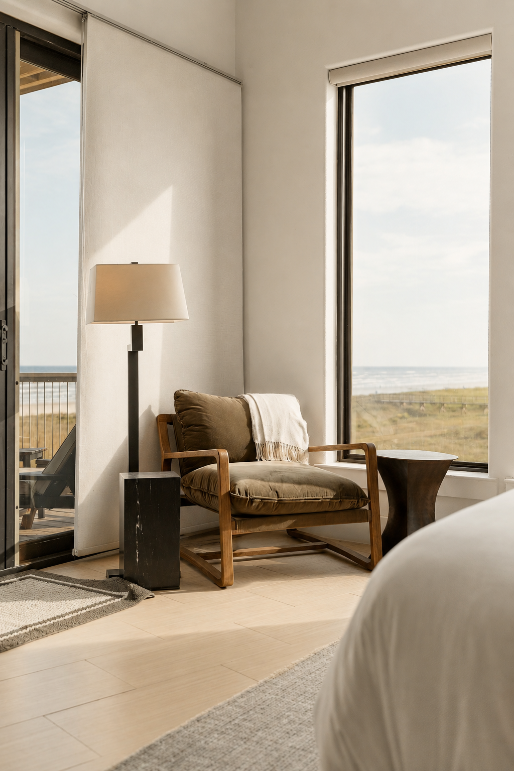Living room with a wooden armchair, a beige cushion, and a white throw blanket near a large window showing an outdoor view of a beach and grassy landscape, with a black side table and floor lamp.