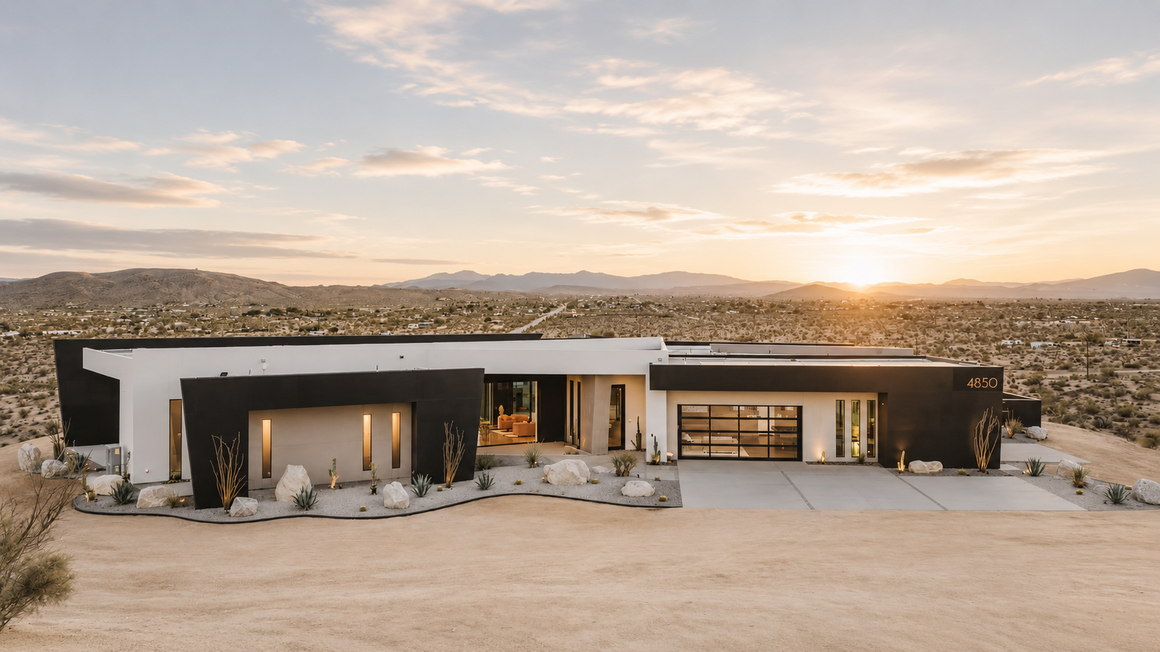 Modern house in desert landscape during sunset, with a black and white exterior, large windows, and a front yard with desert plants and rocks.