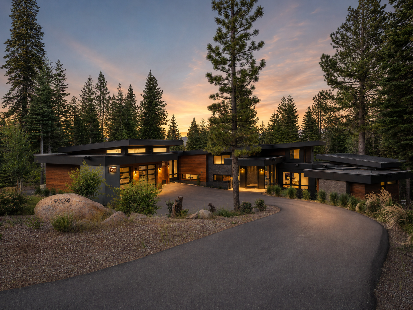 Modern house with large windows and a flat roof, situated in a forested area with tall pine trees and a winding driveway at sunset.