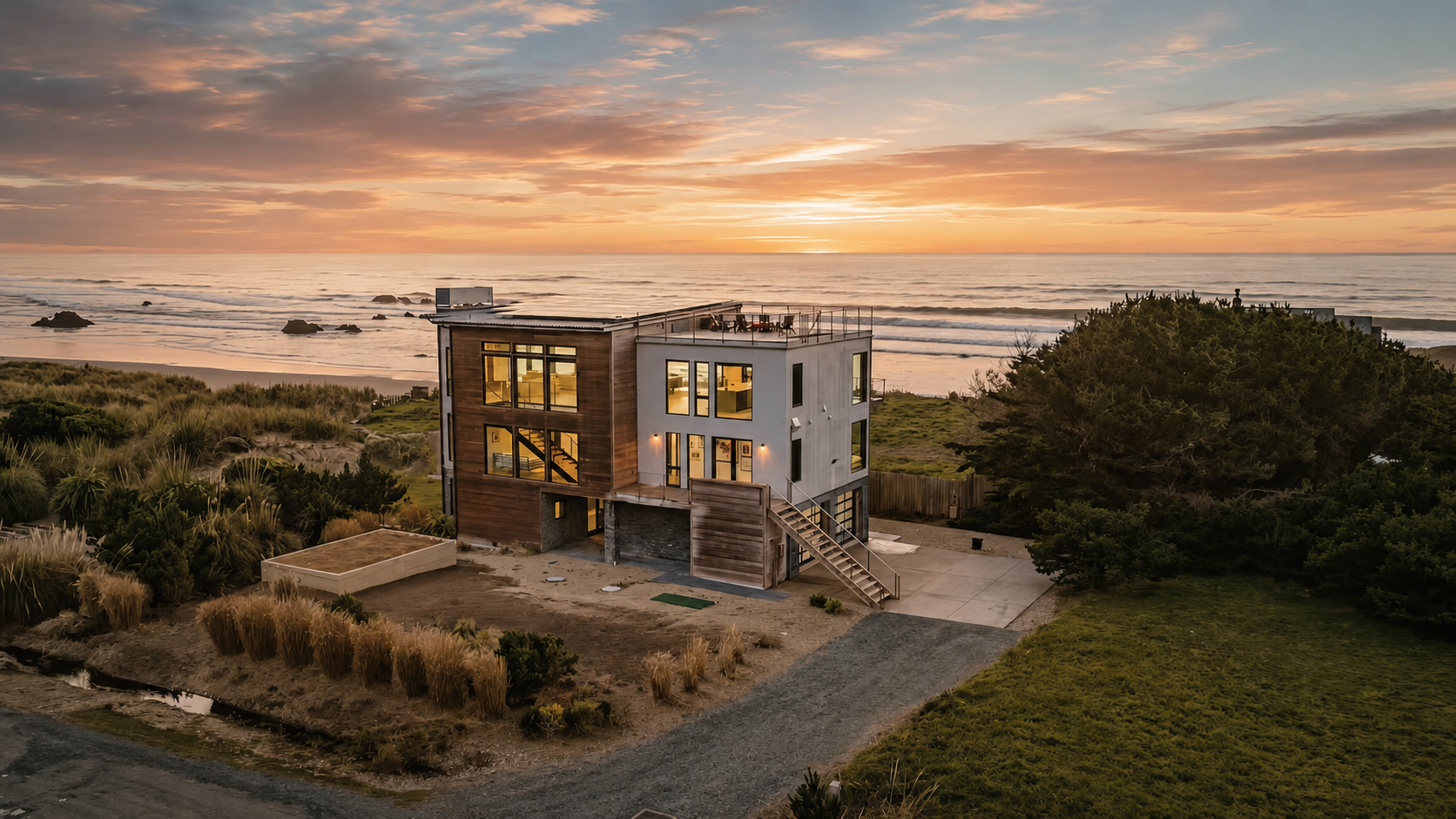 Modern house on the beach at sunset with ocean in the background, large windows, and surrounding greenery.