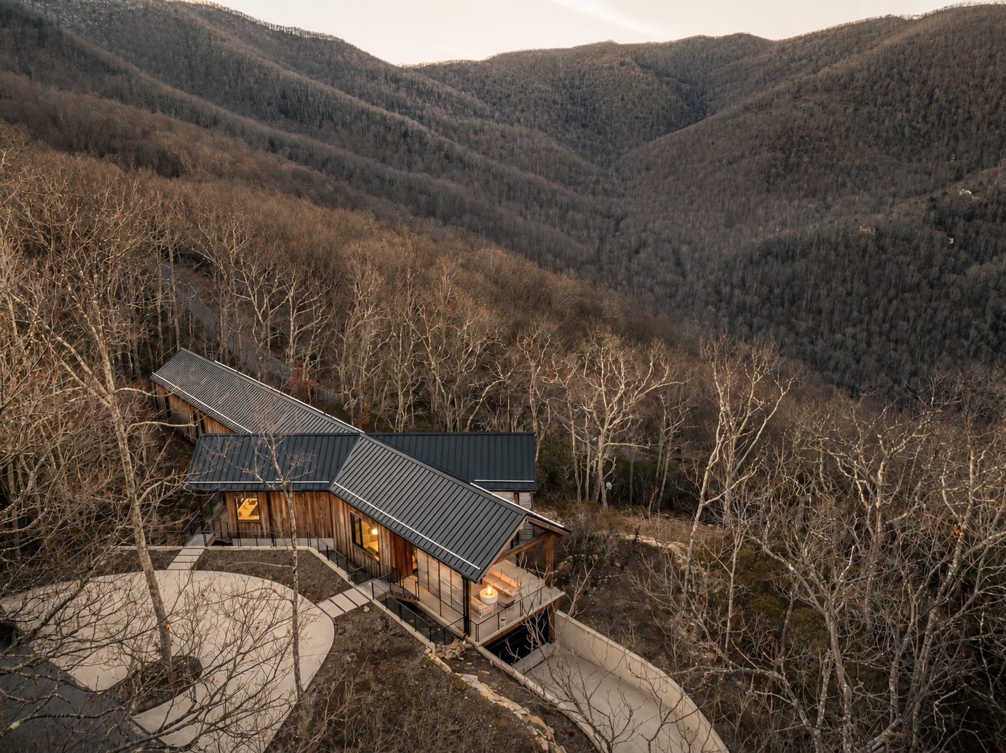 A house with a modern design and metal roof in a wooded mountainous area during late fall or winter, with no leaves on the trees, illuminated by warm interior lighting.