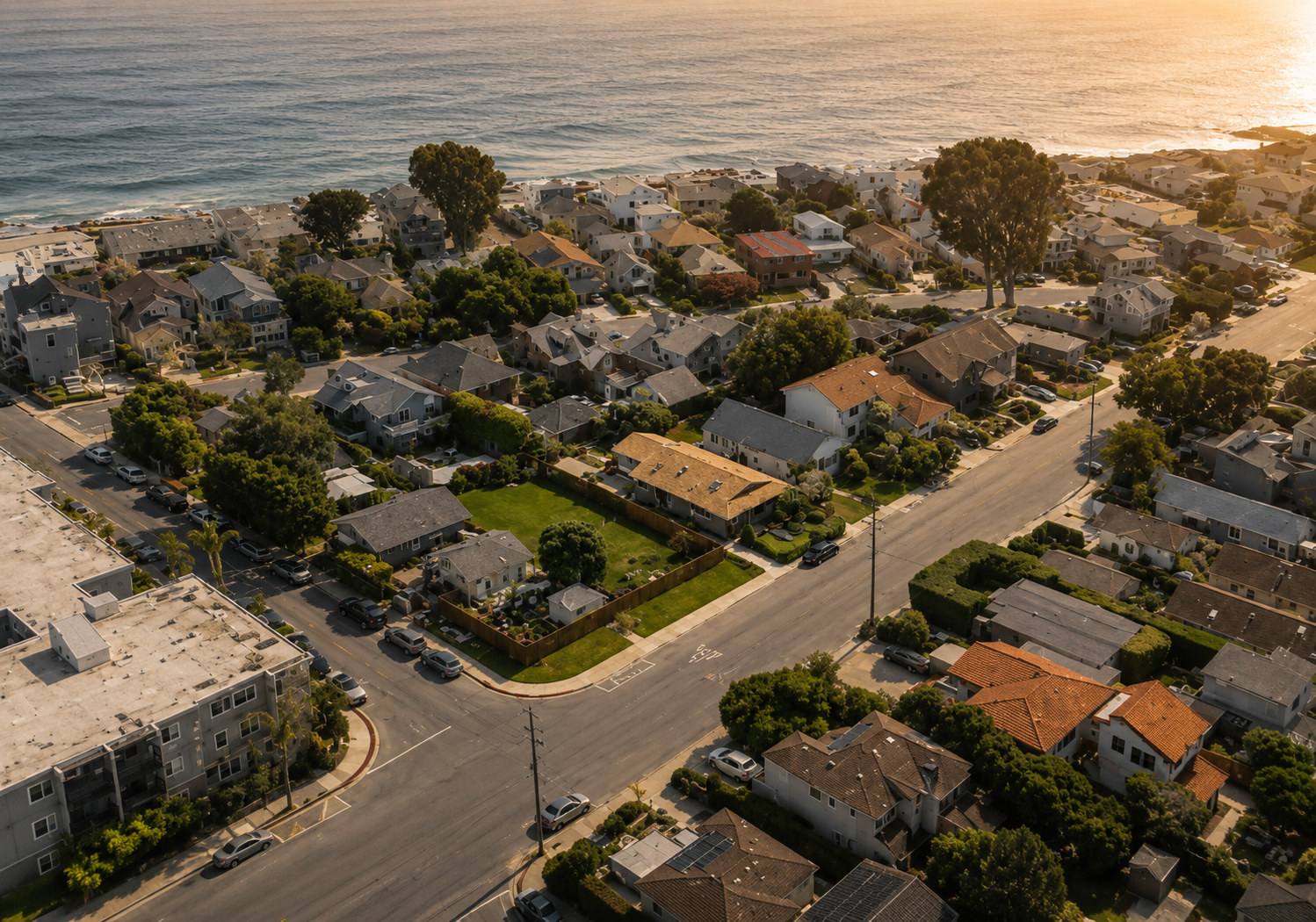 Aerial view of a coastal neighborhood with houses, trees, streets, and the ocean in the background during sunset.