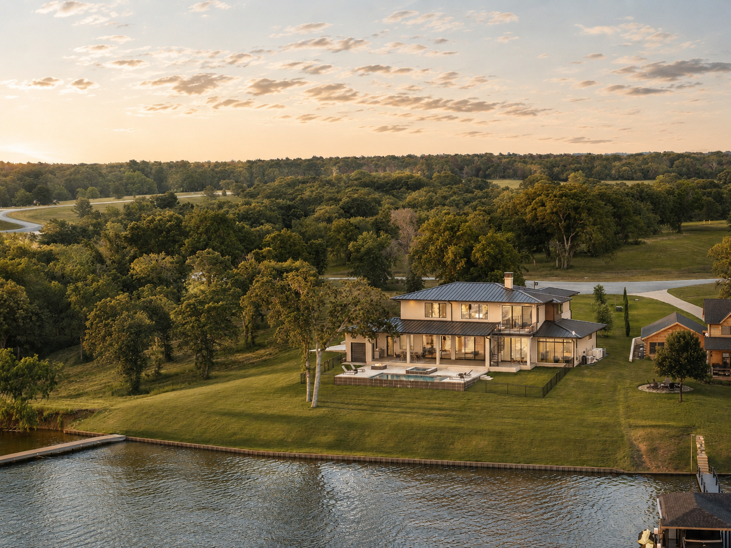 A modern house with large windows and a metal roof situated on a grassy hill next to a body of water, with trees and a scenic landscape in the background during sunset.