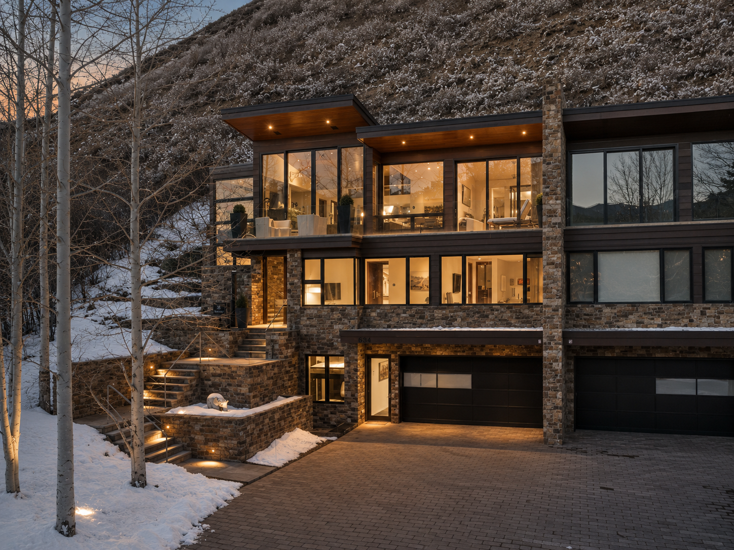 Modern multi-story house with large glass windows and stone exterior, situated on a snowy landscape in the evening