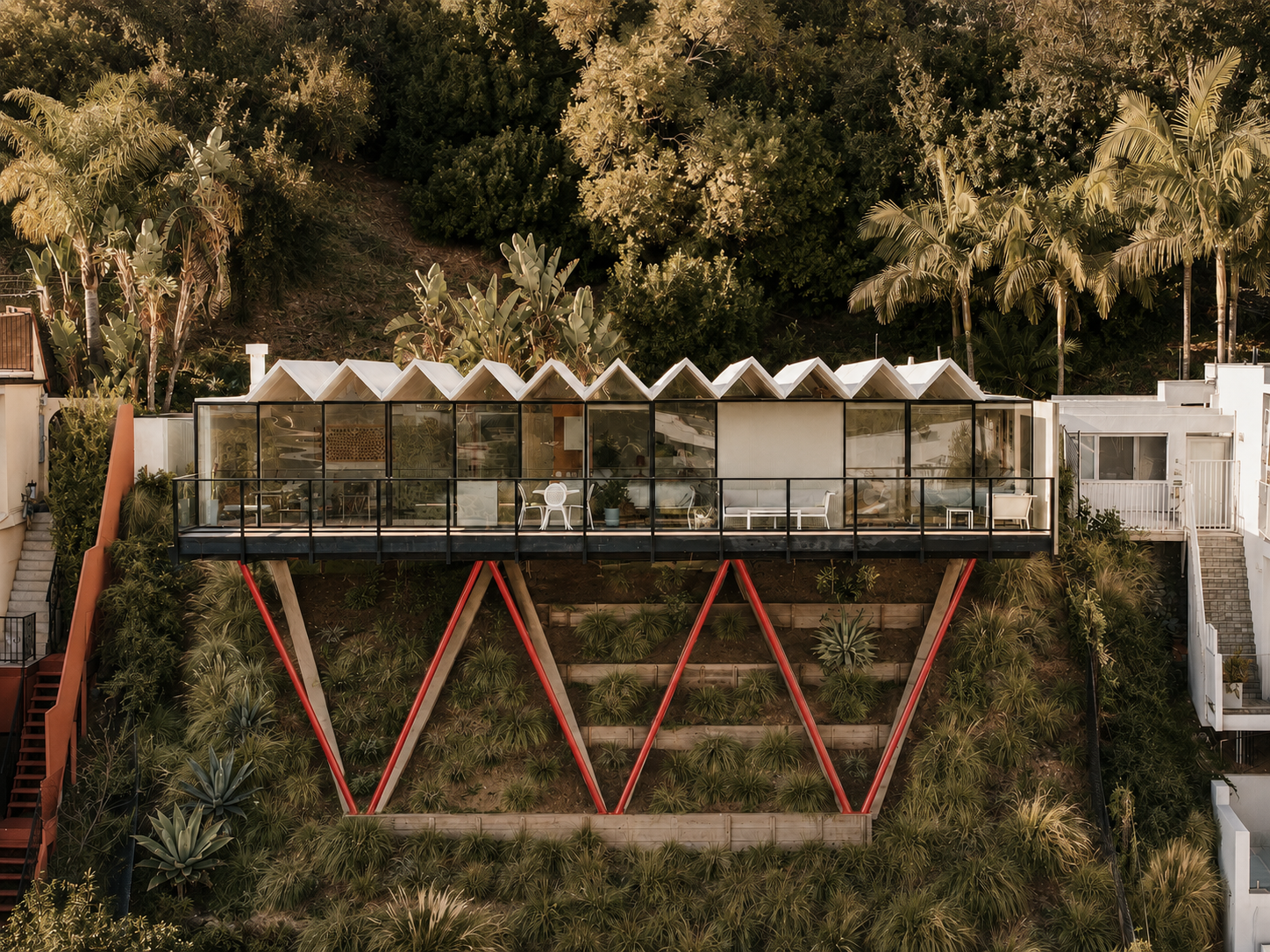 Modern house built on a hillside with large glass windows and a red A-frame support structure, surrounded by greenery and palm trees.