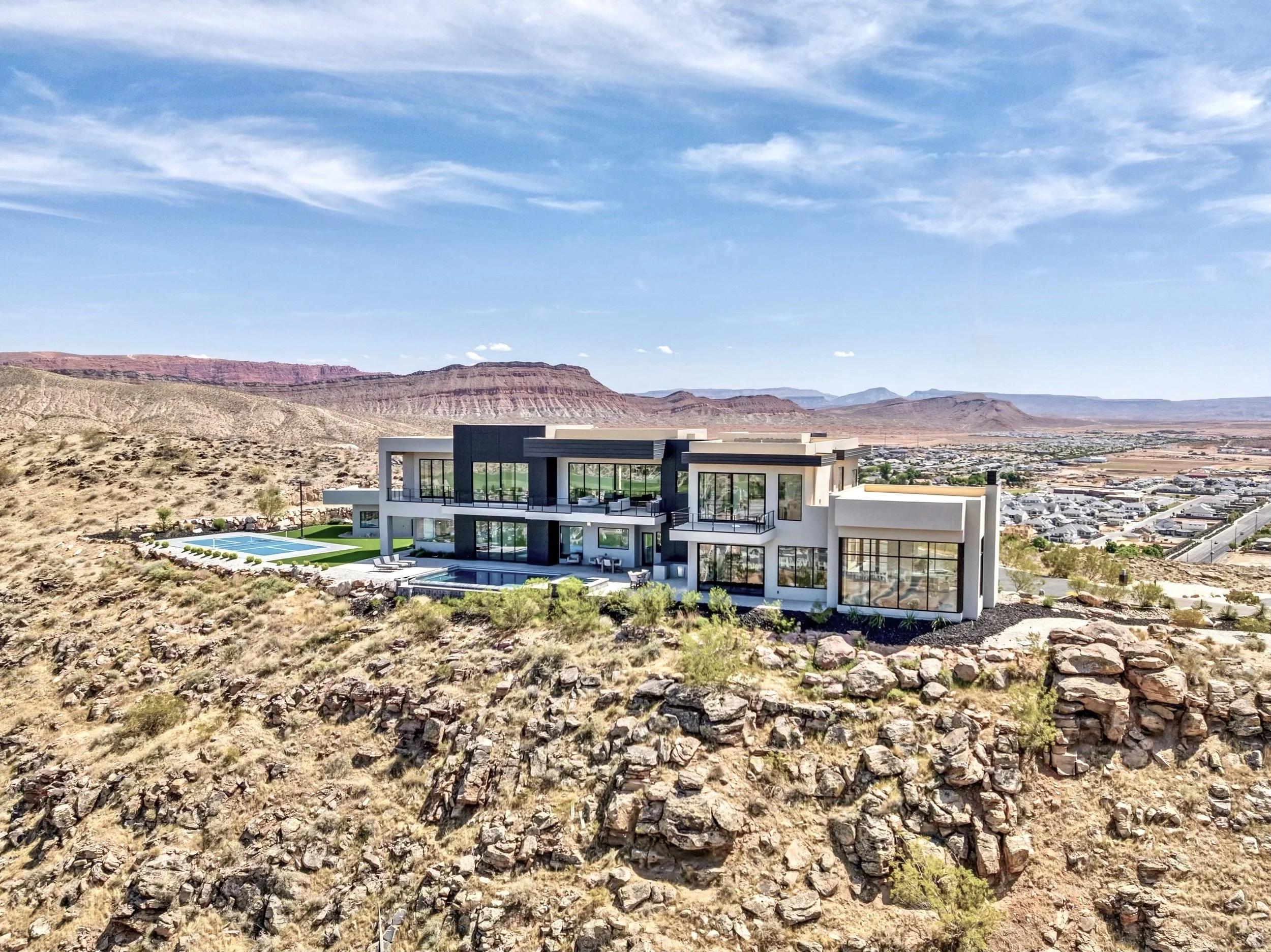 A modern house with large glass windows and a swimming pool, set against a desert landscape with mountains and a blue sky with scattered clouds.