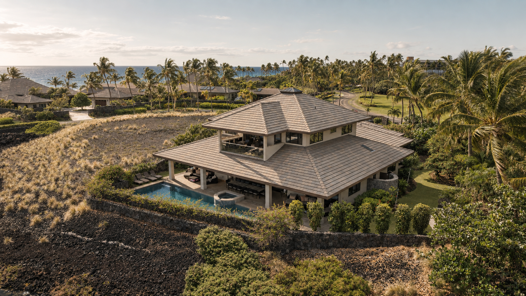 Aerial view of a large house with a pool, surrounded by tropical trees and a coastal landscape with the ocean in the background.