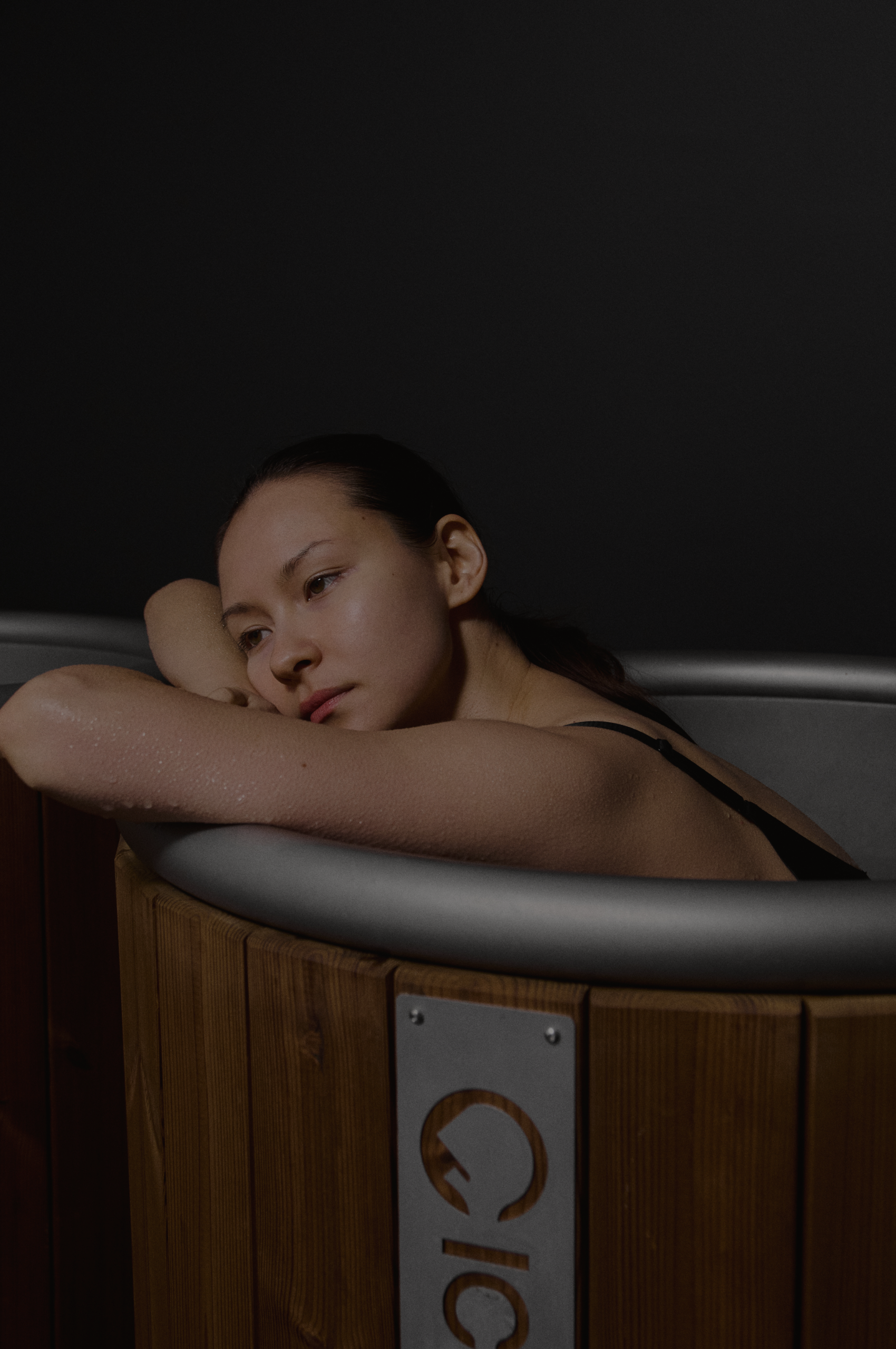 A woman resting her head on her arm inside a wooden hot tub or sauna, with a contemplative expression.