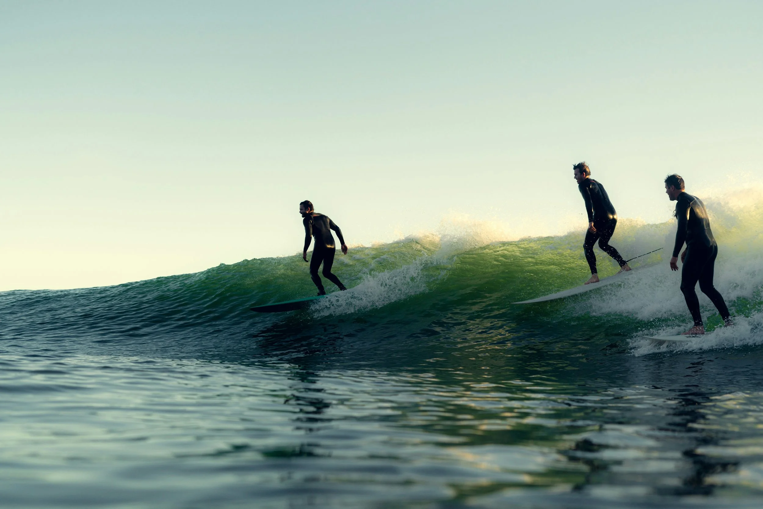 Trois personnes en combinaison de plongée pratiquent le surf sur une vague dans l'océan lors d'une journée ensoleillée.