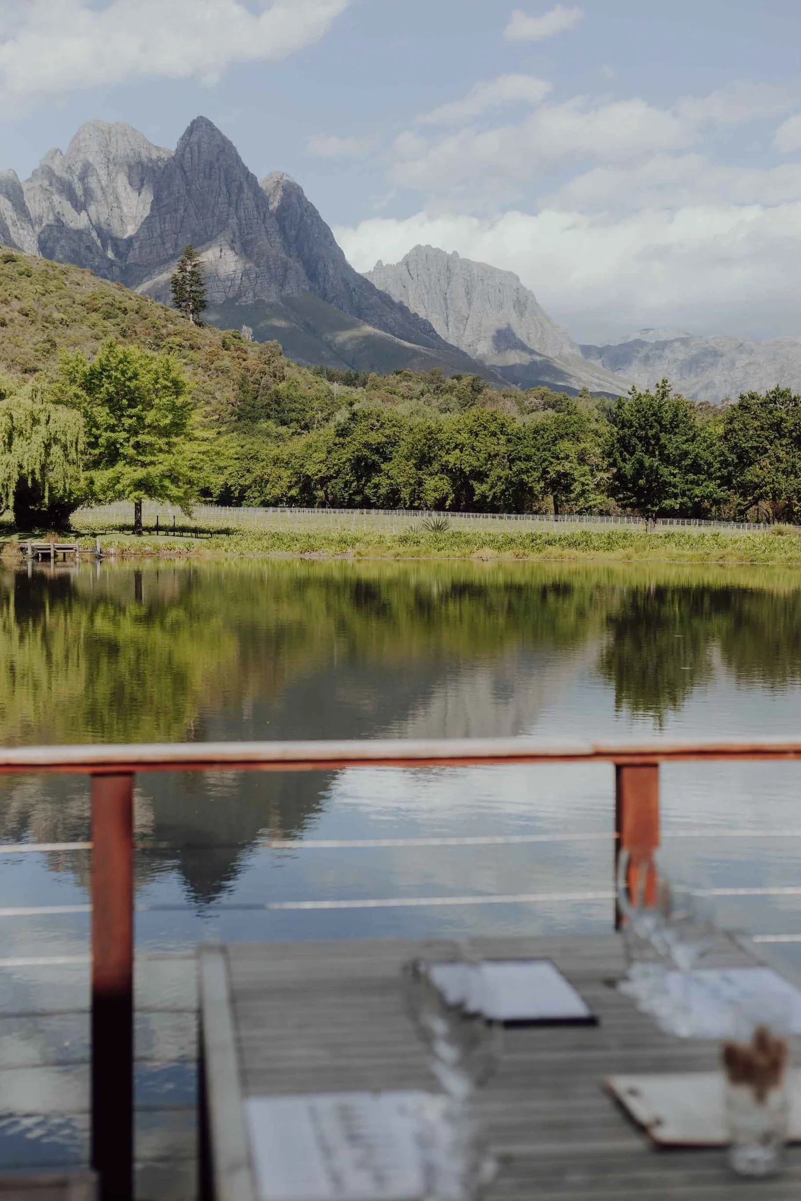 A scenic view of a mountain landscape with a lake in the foreground, trees, and mountain peaks under a partly cloudy sky.