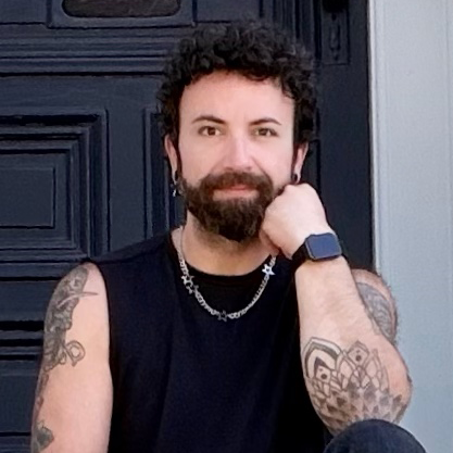 Stylist (Christian) with curly hair and beard, wearing a black sleeveless shirt, silver chain necklace, and smartwatch, sitting in front of a black garage door.