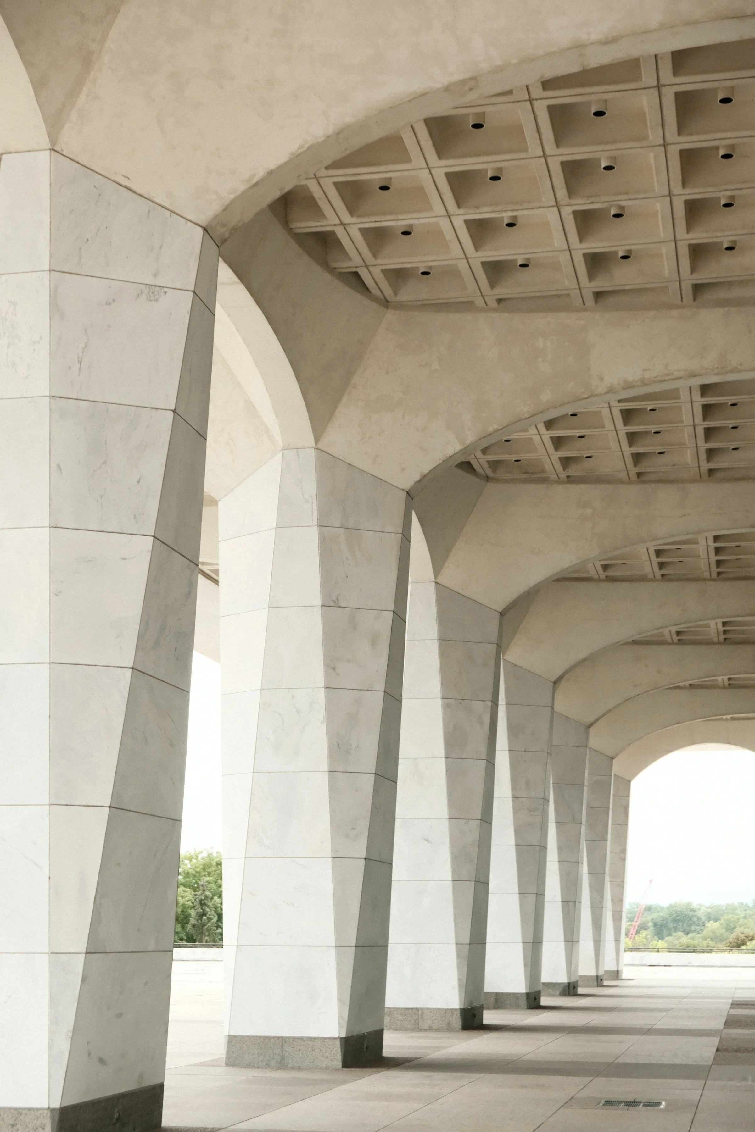 A row of tall, geometric, white stone columns supporting a curved ceiling with square recesses and small circular openings.