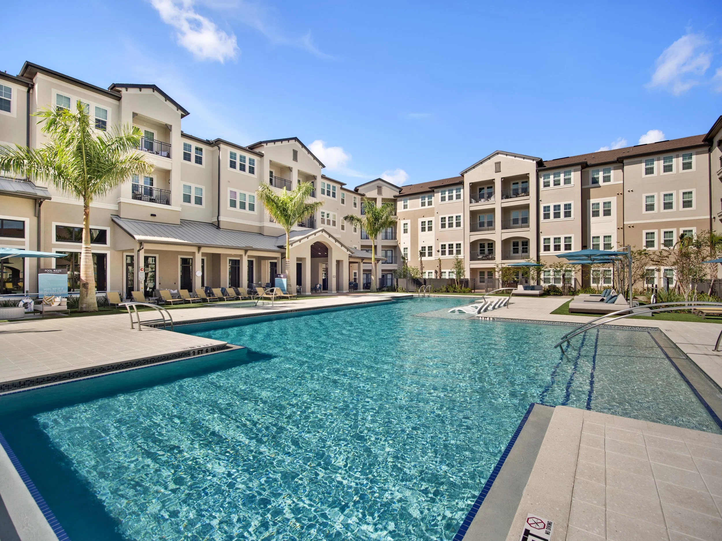 Apartment complex with a swimming pool, lounge chairs, umbrellas, and palm trees under a blue sky.