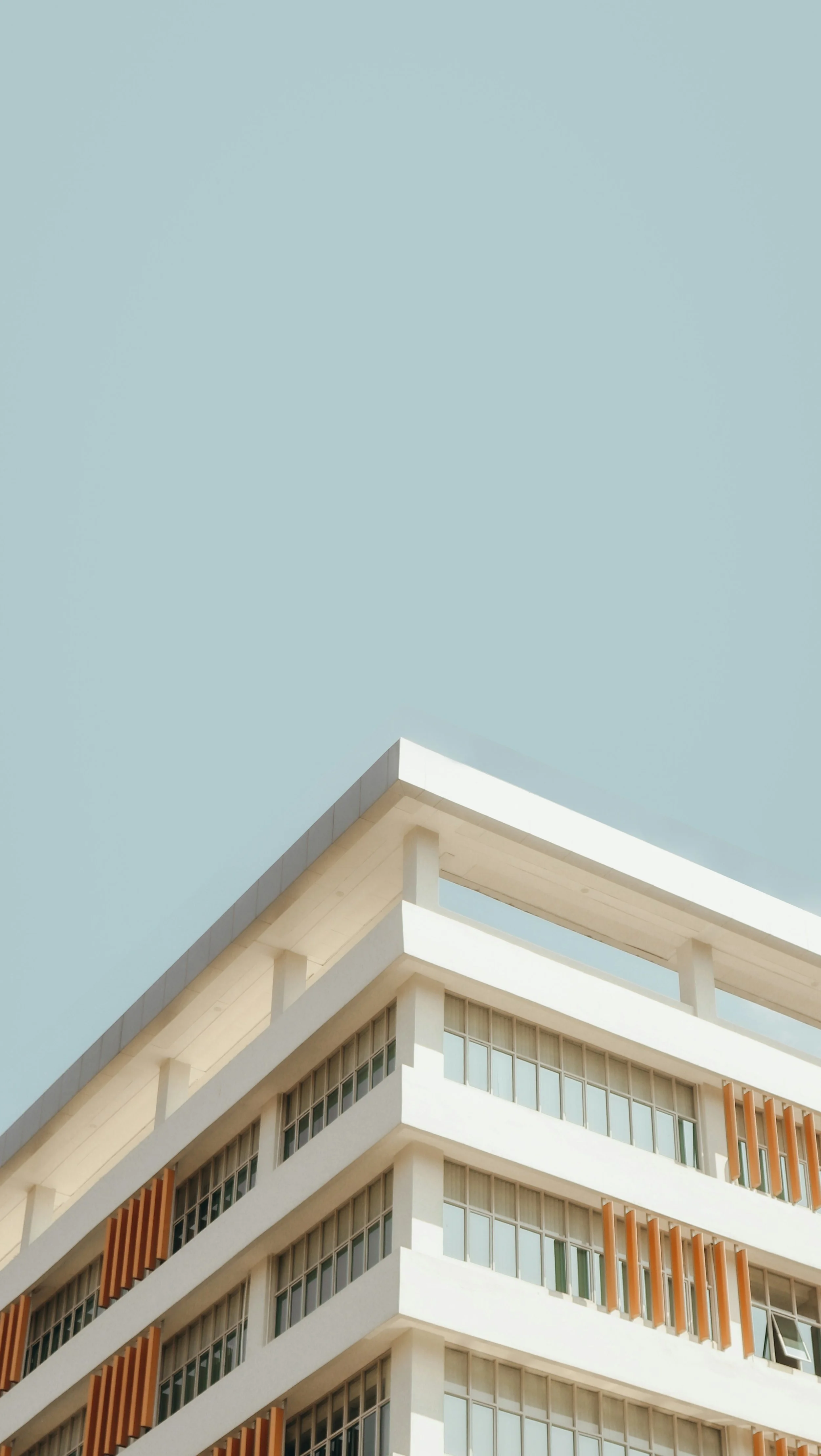 Modern white multi-story building with large windows and orange slats, against a light blue sky.