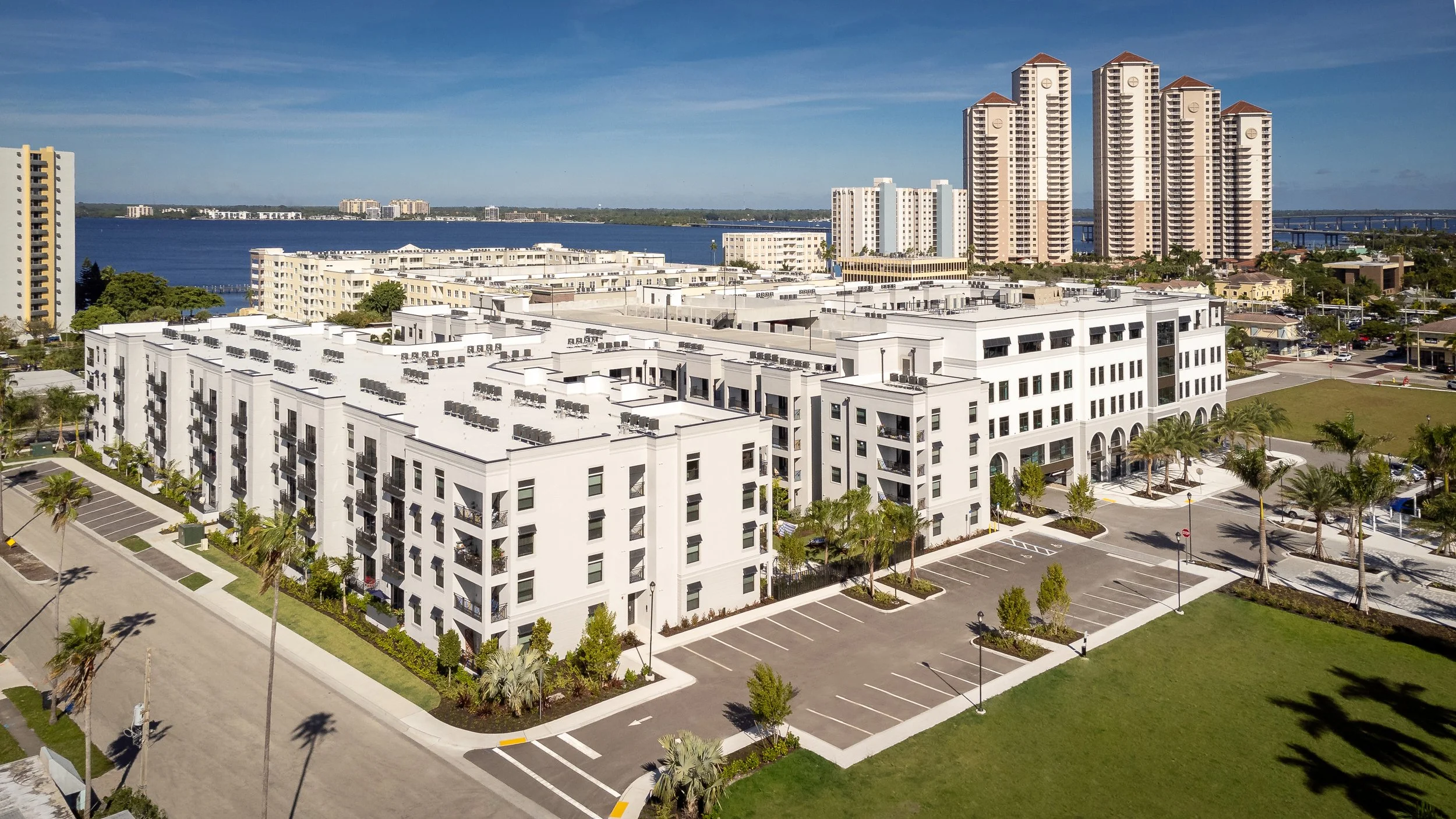 Aerial view of modern white residential buildings with parking lots surrounded by palm trees, with high-rise buildings and a body of water in the background under a blue sky.