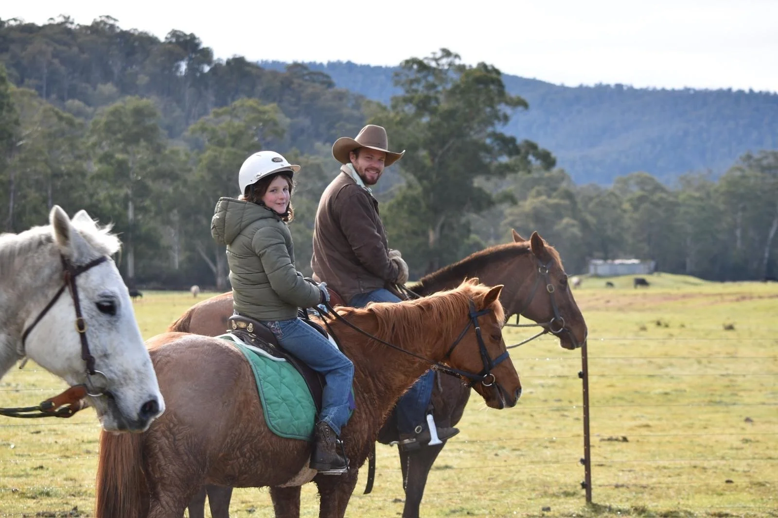 A man and a girl riding horses in a grassy field with a wooded hillside in the background.