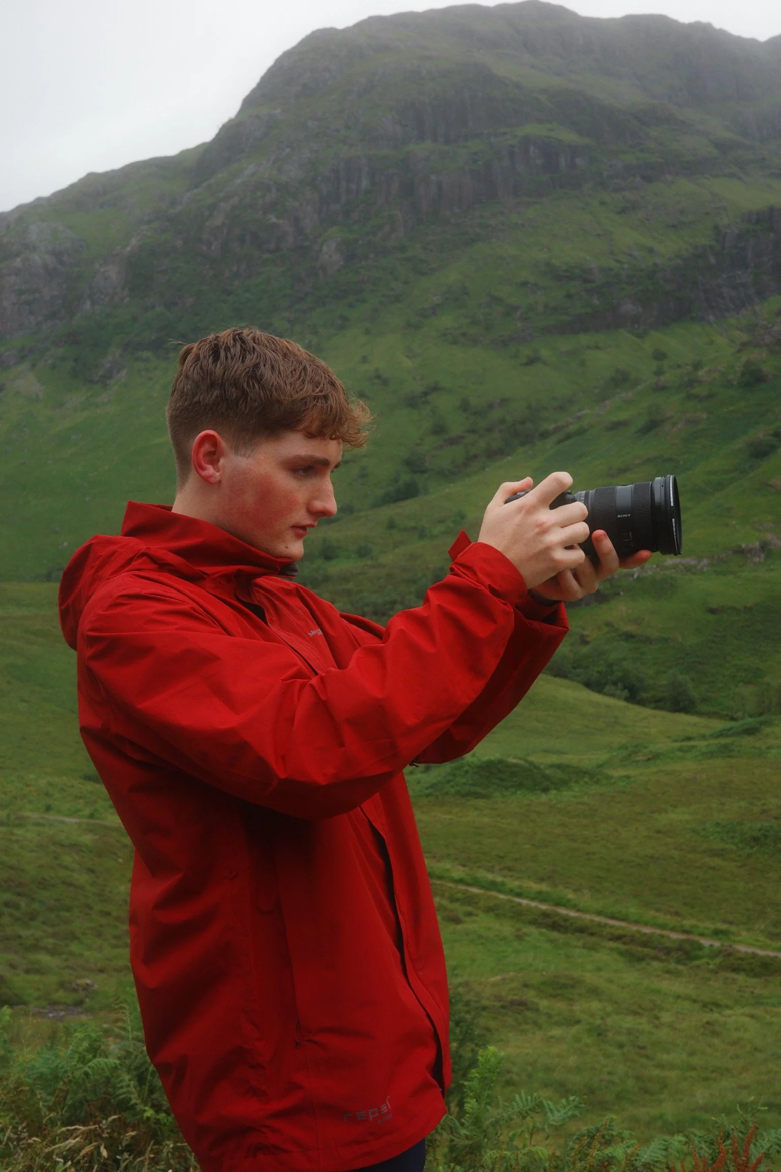 A young man with brown hair in a red jacket holding a camera outdoors in front of green rolling hills and a large mountain.