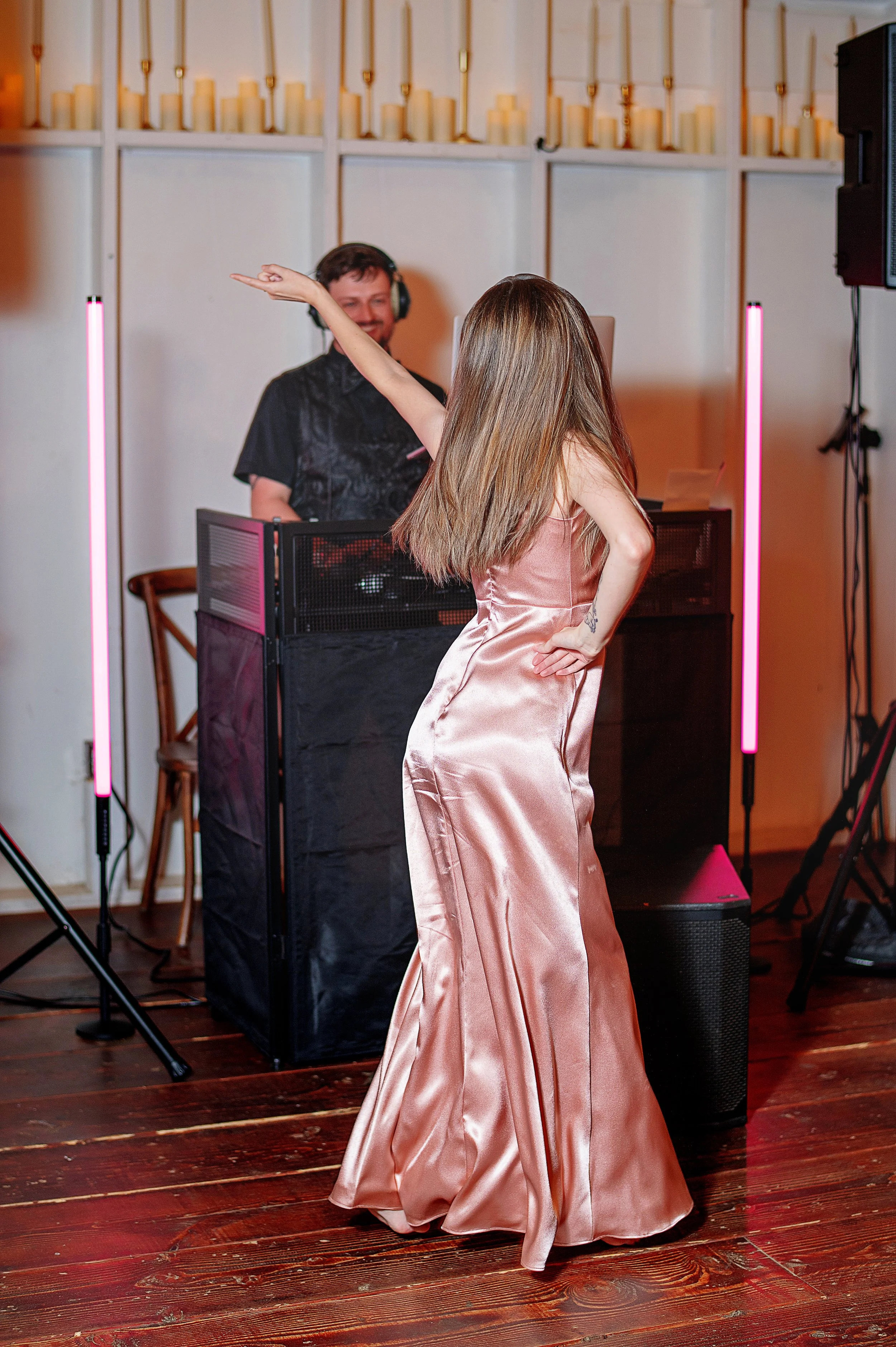 A woman in a silky pink gown dances with her back to the camera while a man at a DJ booth watches her, smiling, in a warmly lit room decorated with candles and pink neon lights.