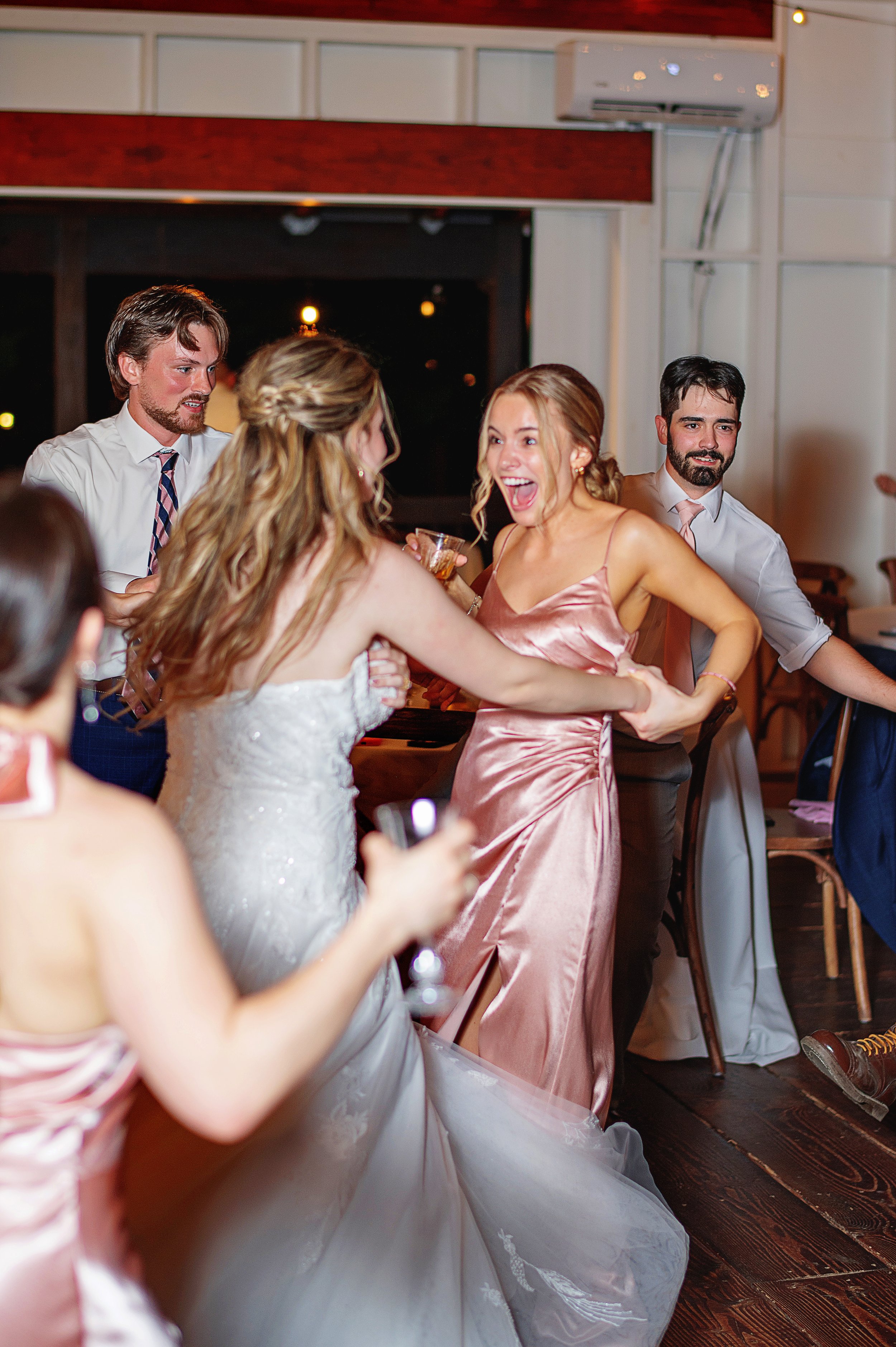People at a party or celebration, with women in dresses and men in shirts, engaging in conversation and holding drinks, in a cozy indoor setting with wood floors and a window in the background.