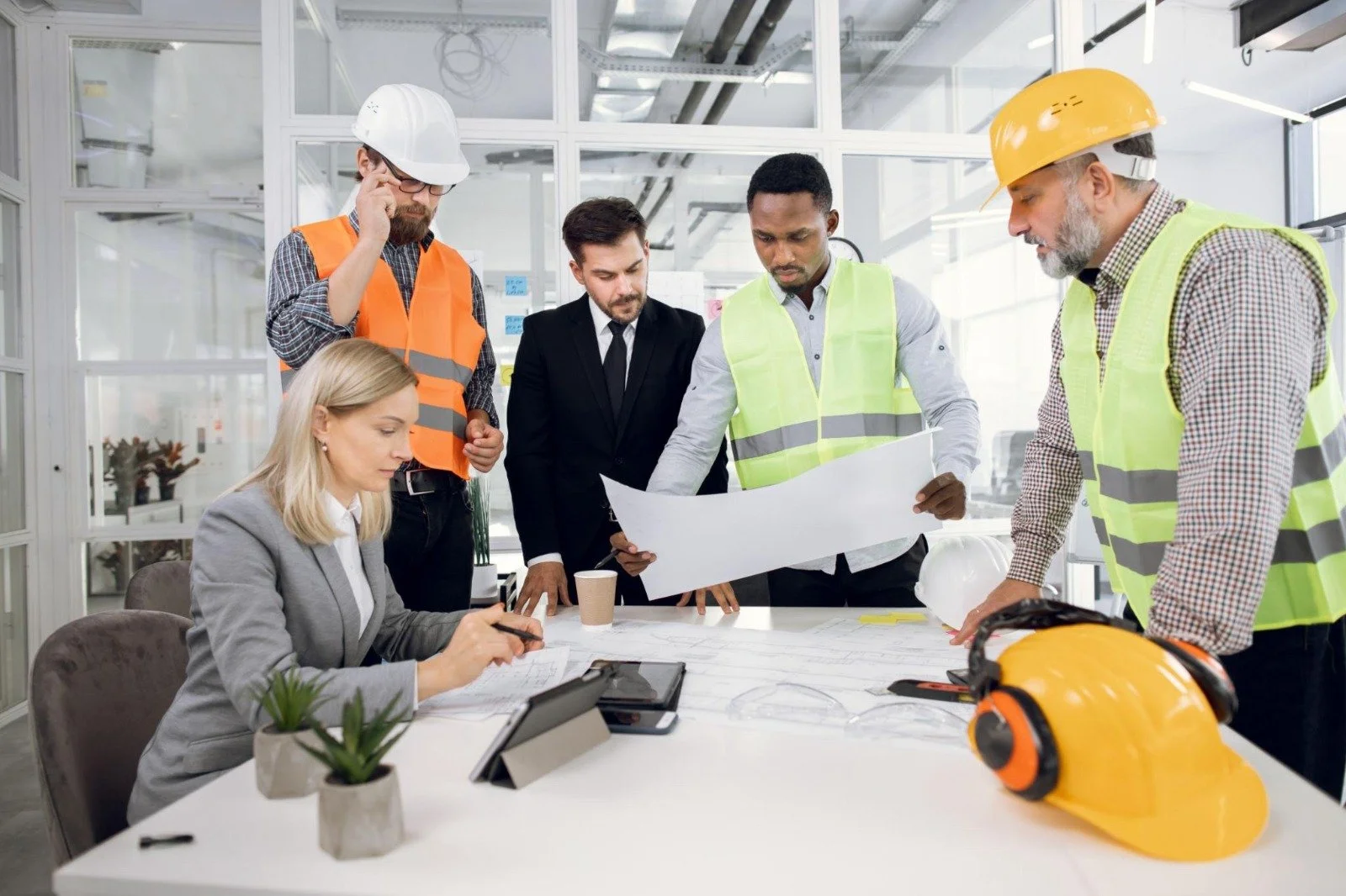 Group of construction professionals reviewing blueprints in an office setting. One woman is seated taking notes, while four men, some in safety helmets and vests, are standing discussing plans.