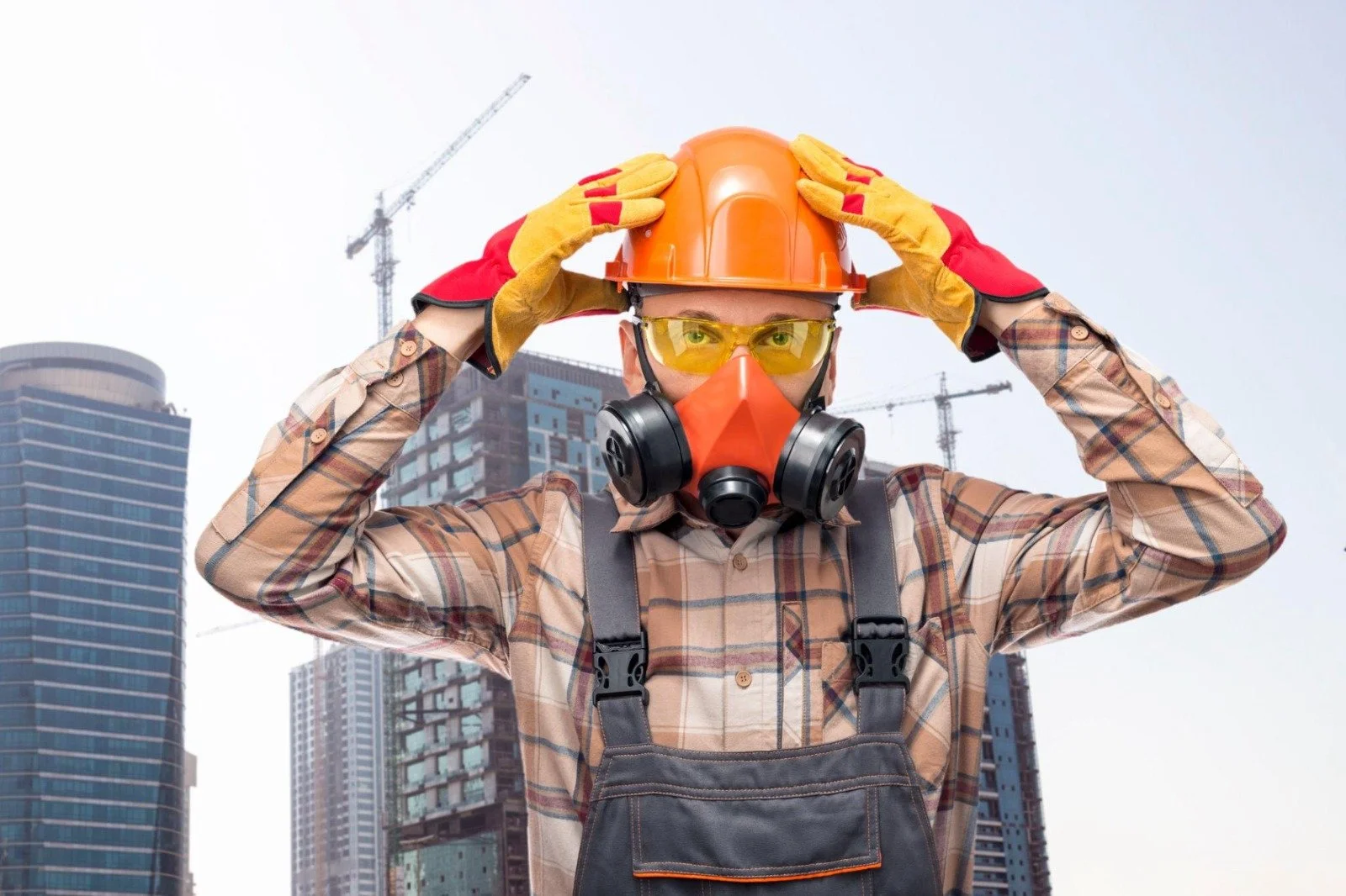 A construction worker in safety gear, including a yellow hard hat, yellow safety glasses, a respirator mask, and gloves, standing in front of a cityscape with tall buildings and cranes.