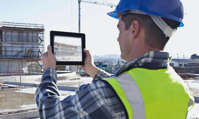 Construction worker taking a photo of the building site with a tablet, wearing a blue safety helmet and a yellow safety vest.
