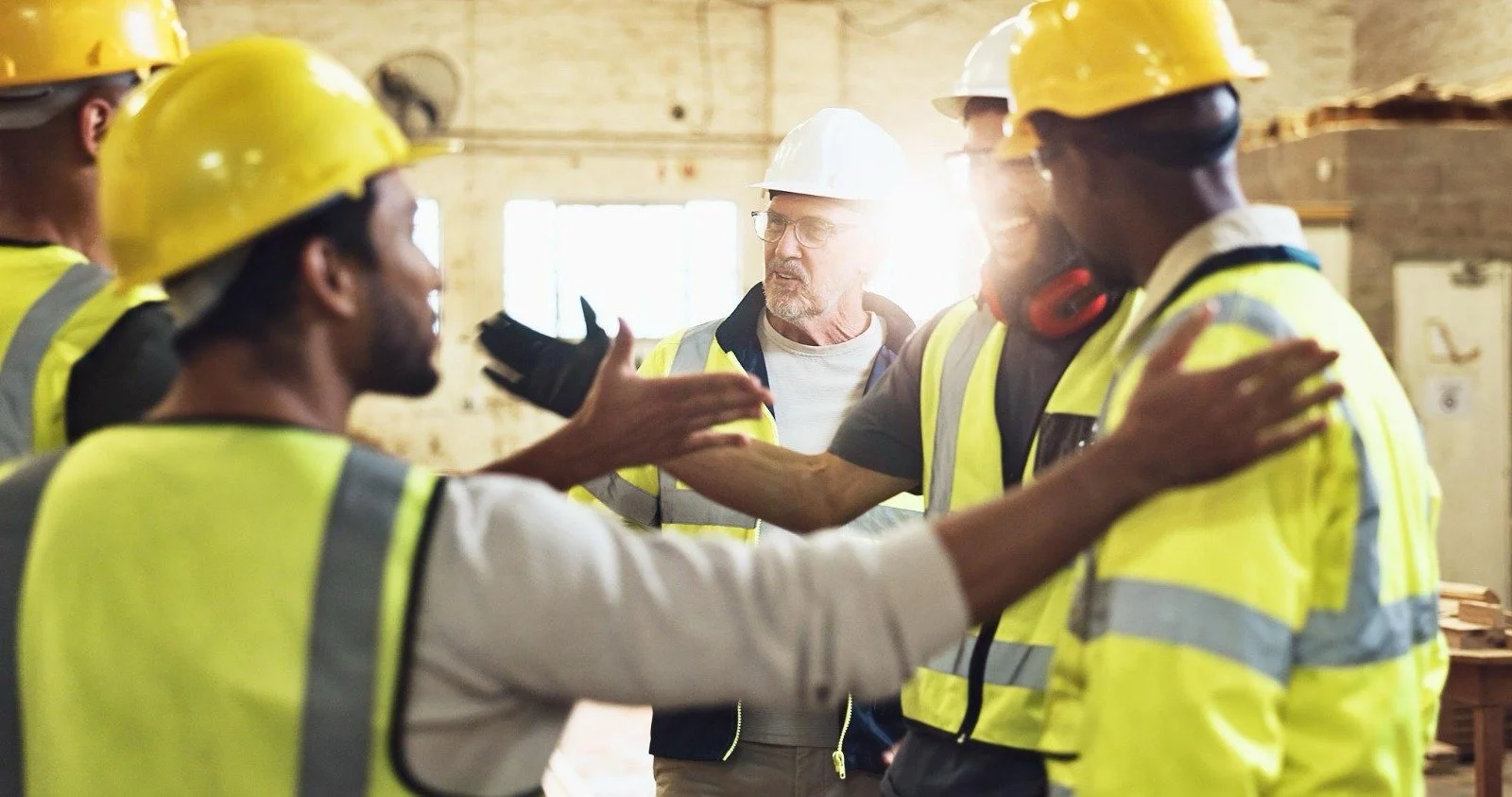 Workers in safety vests and helmets having a discussion in a construction site or warehouse. One person is gesturing with their hand while others listen.