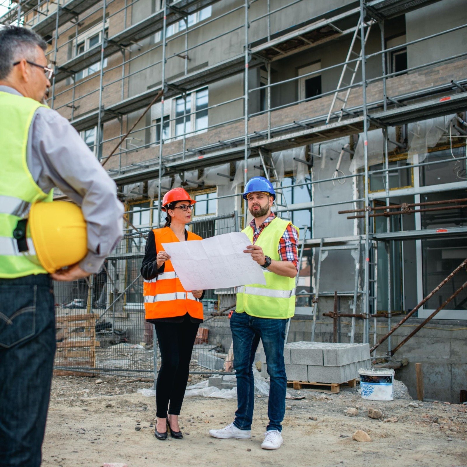 Construction site with four professionals, including two women and two men, wearing safety helmets and vests, discussing plans. An unfinished building with scaffolding is in the background.