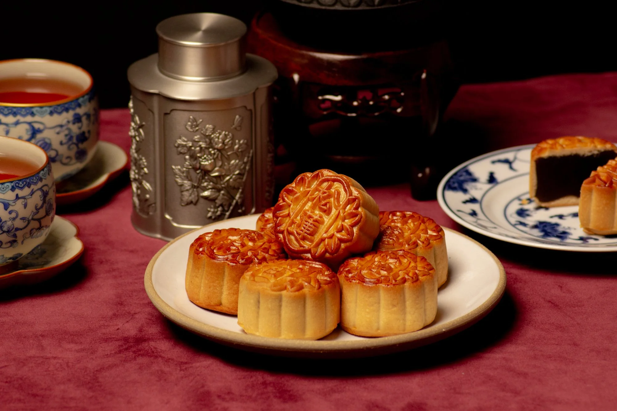 Mooncakes on a white plate with tea cups, a metal tea canister, and a blue and white patterned plate with mooncake slices on a red fabric background.