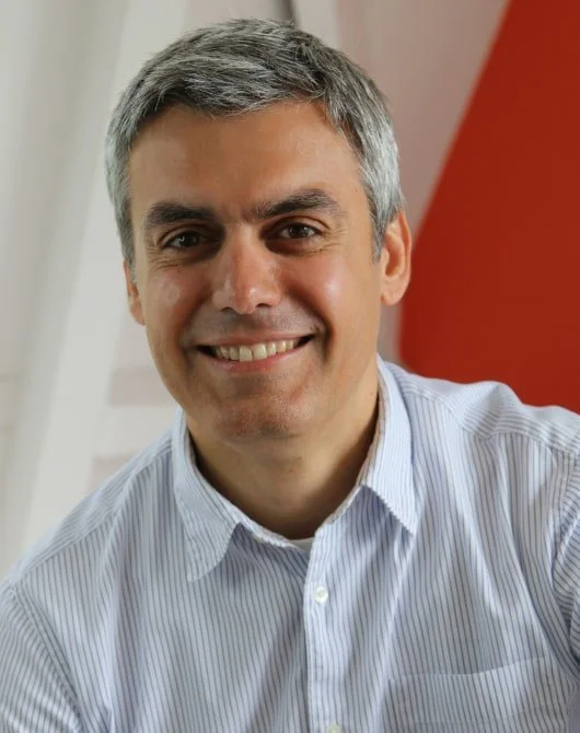Smiling middle-aged man with gray hair wearing a light blue, striped collared shirt, indoors with a red wall background.
