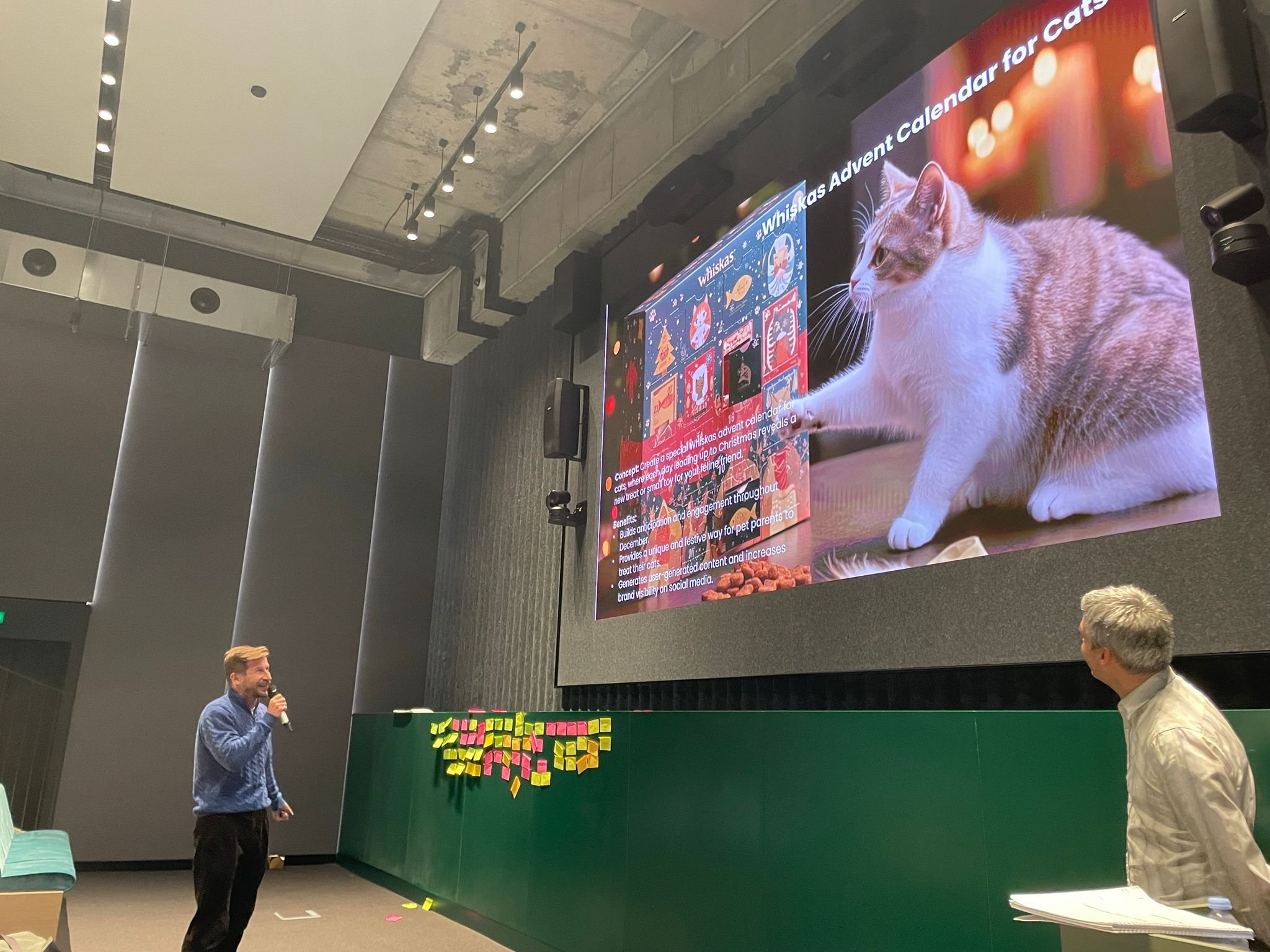 A presentation in a conference room showing a large screen with a Whiskas Advent Calendar for Cats, featuring a picture of a cat and holiday-themed decorations. Two men are standing in front of the screen, one speaking into a microphone and the other looking at the screen.