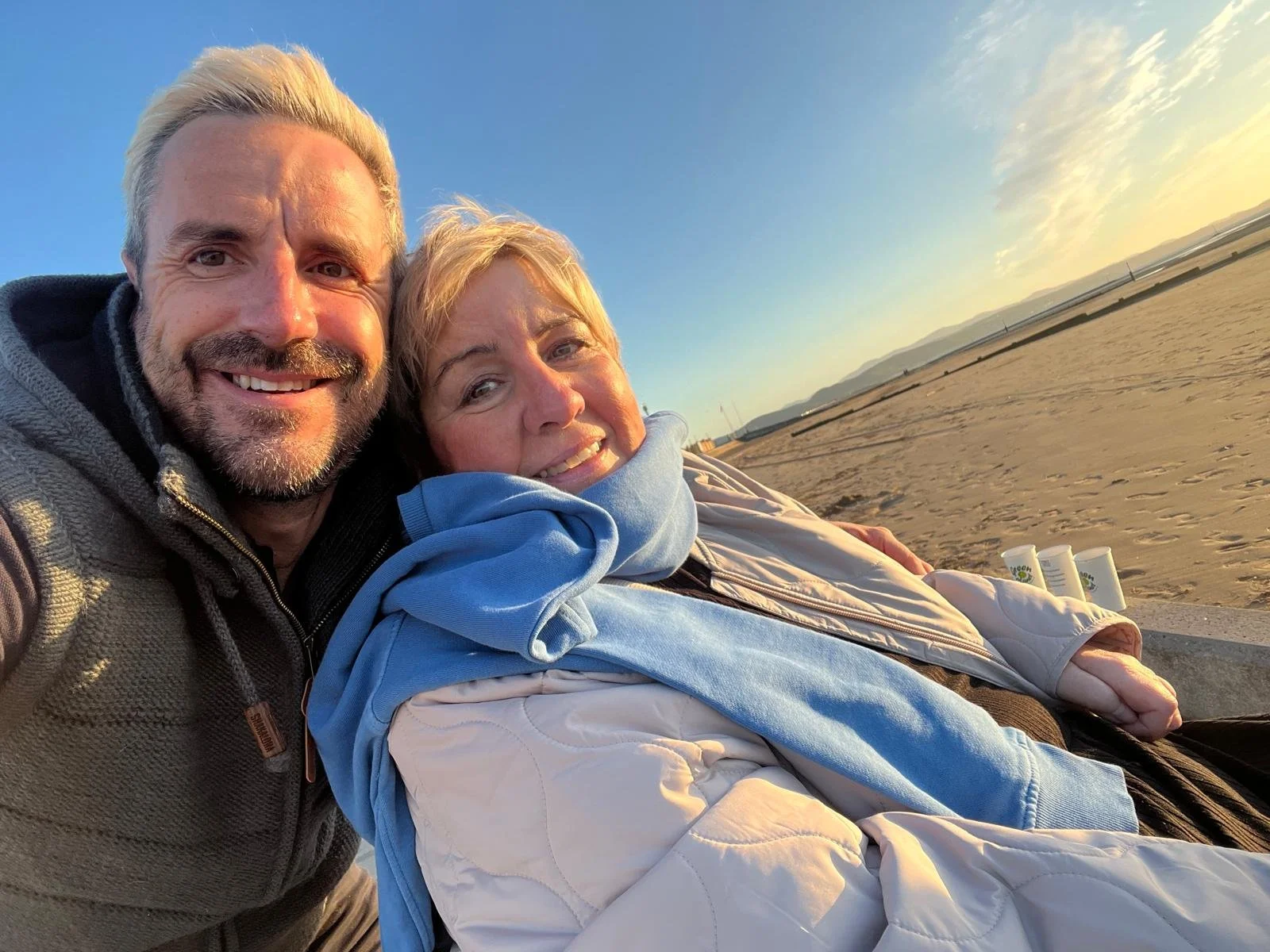 A smiling middle-aged man and woman taking a selfie on a beach during sunset, with the ocean and mountains in the background.