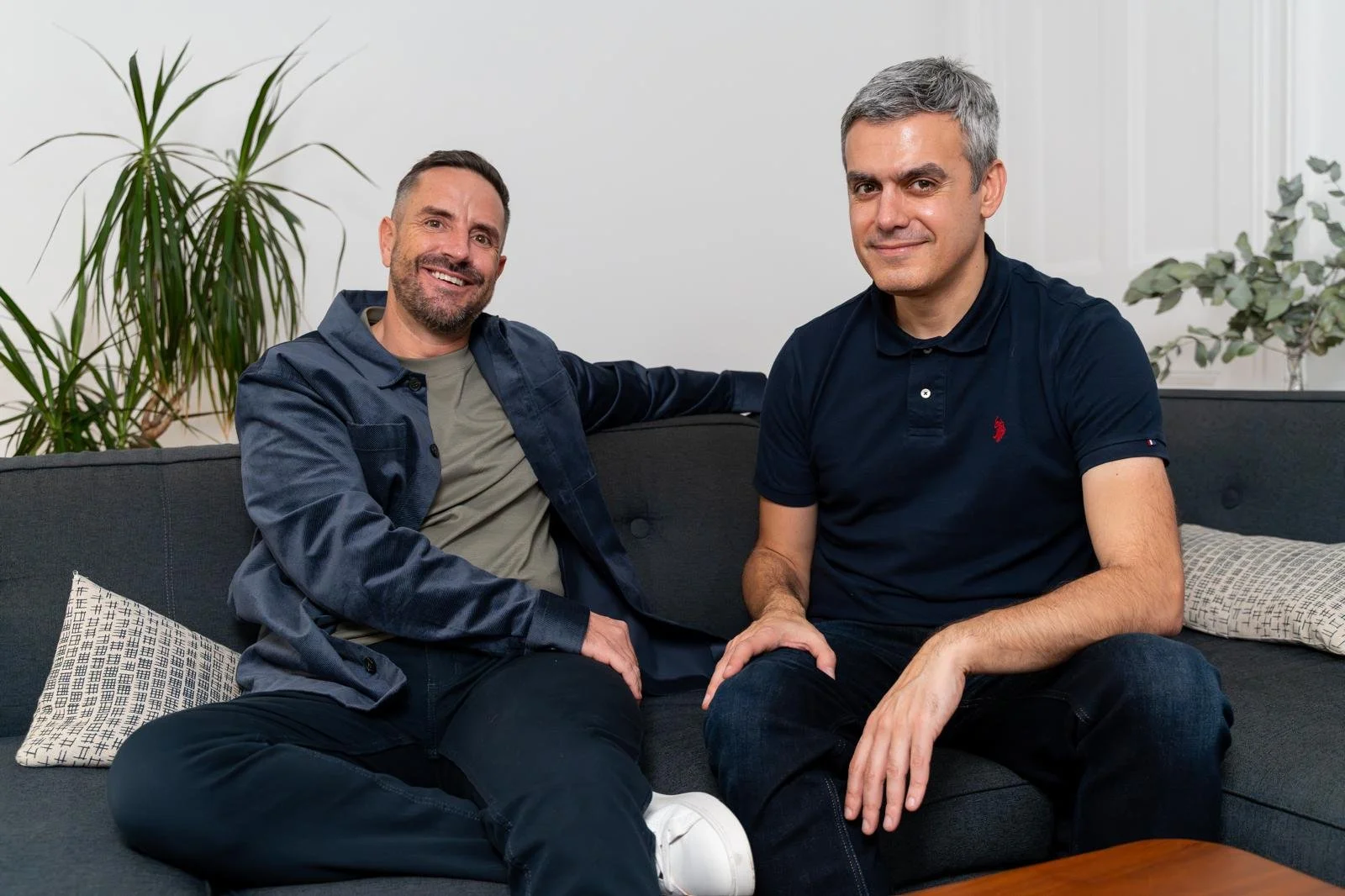 Two men sitting on a gray couch in a living room, smiling at the camera. One man has a beard and wears a dark jacket over a gray shirt, and the other has gray hair and wears a dark polo shirt. There are green plants behind them and decorative pillows on the couch.