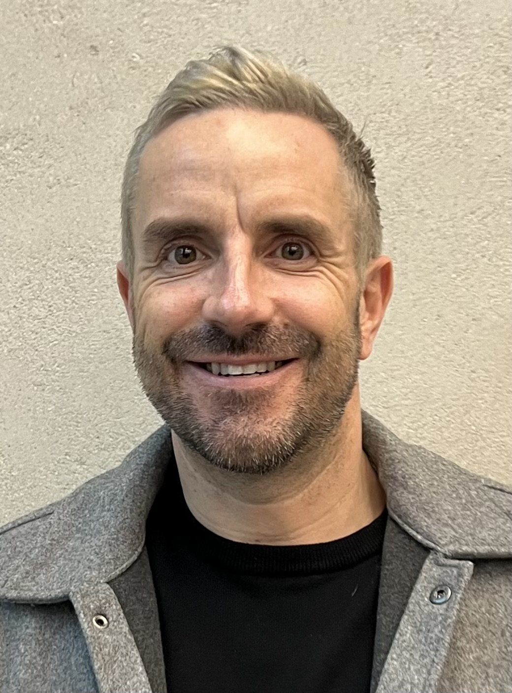 A smiling man with short, light brown hair, a beard, and gray eyes wearing a dark shirt and a gray jacket, standing against a beige textured wall.