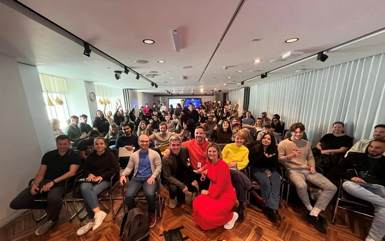 Group of people in a conference room, some sitting and some standing, smiling and looking at the camera, with a presentation on a screen in the background.