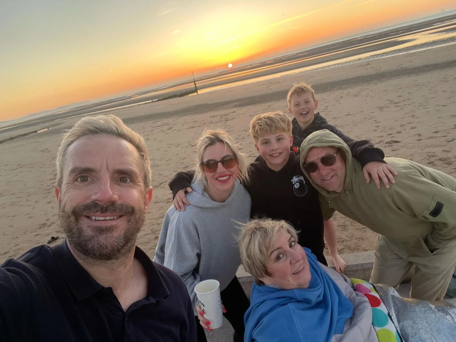 A group of seven people, including children and adults, smiling and posing on a sandy beach during sunset. The sky is orange and yellow, and the ocean is visible in the background.