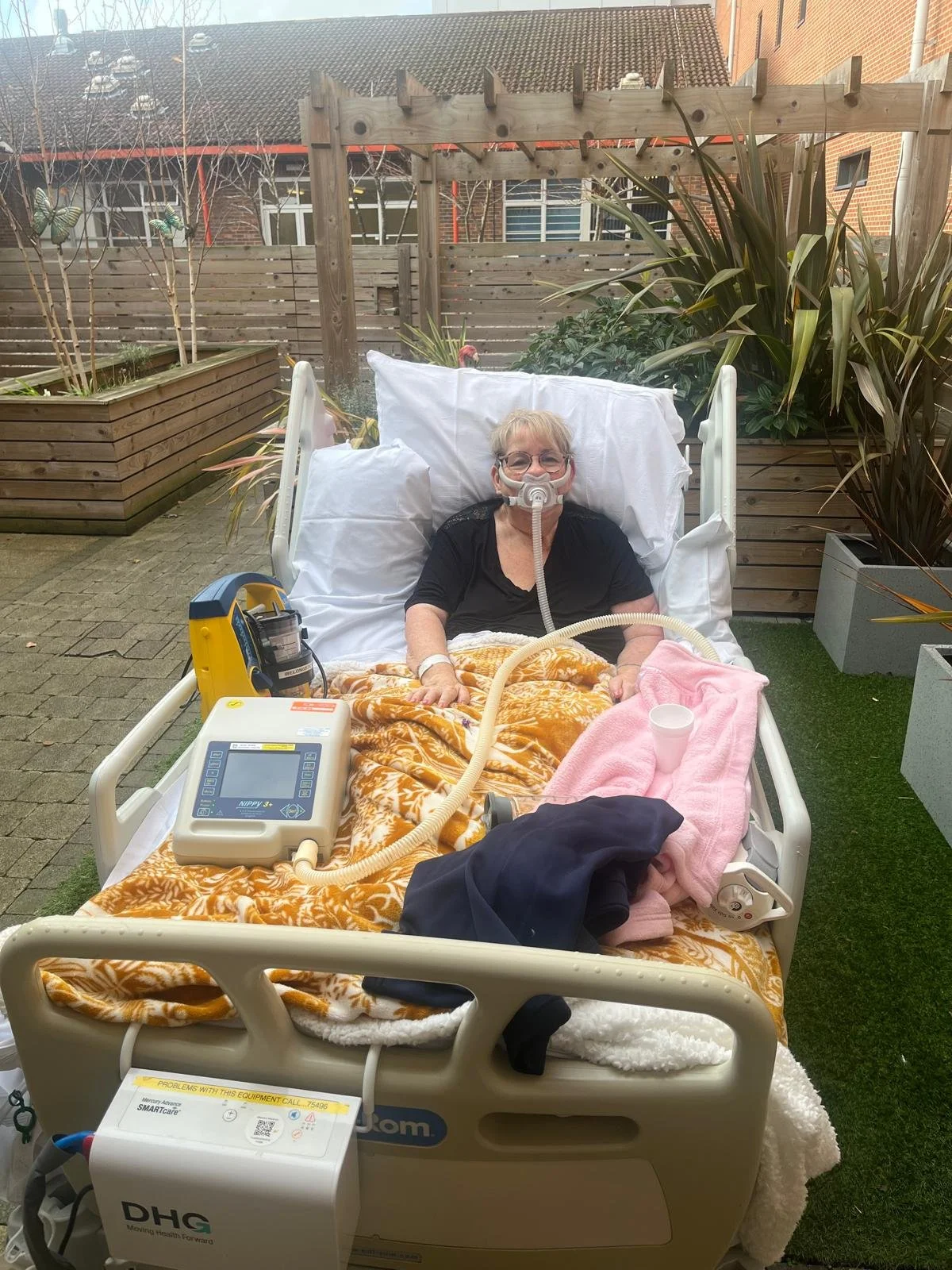 A woman in a hospital bed outdoors, wearing a breathing mask connected to medical equipment, with a blanket covering her. The bed is in a garden area with plants, wooden fencing, and buildings in the background.