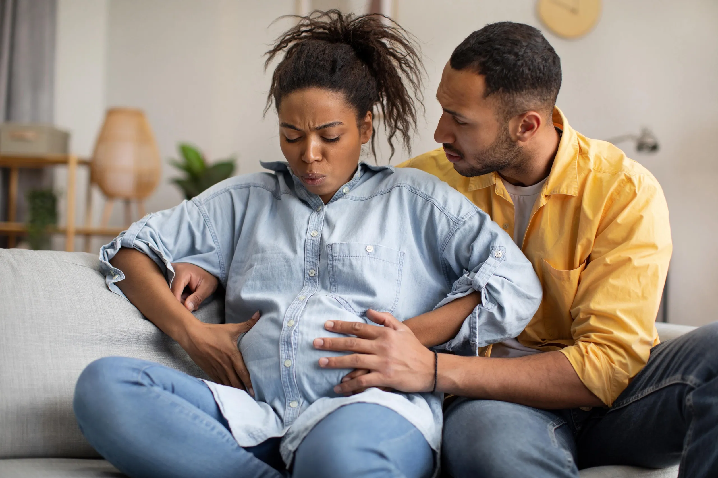 A woman sitting on a sofa experiencing abdominal pain, with a man supporting her and feeling concerned.