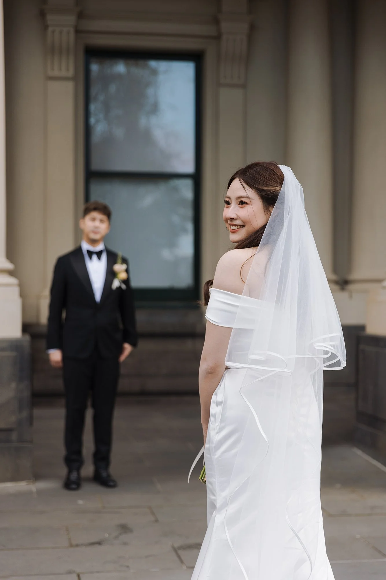A bride in a white off-shoulder wedding dress with a veil, smiling, with a groom in a black tuxedo standing in front of a historic building with large windows.