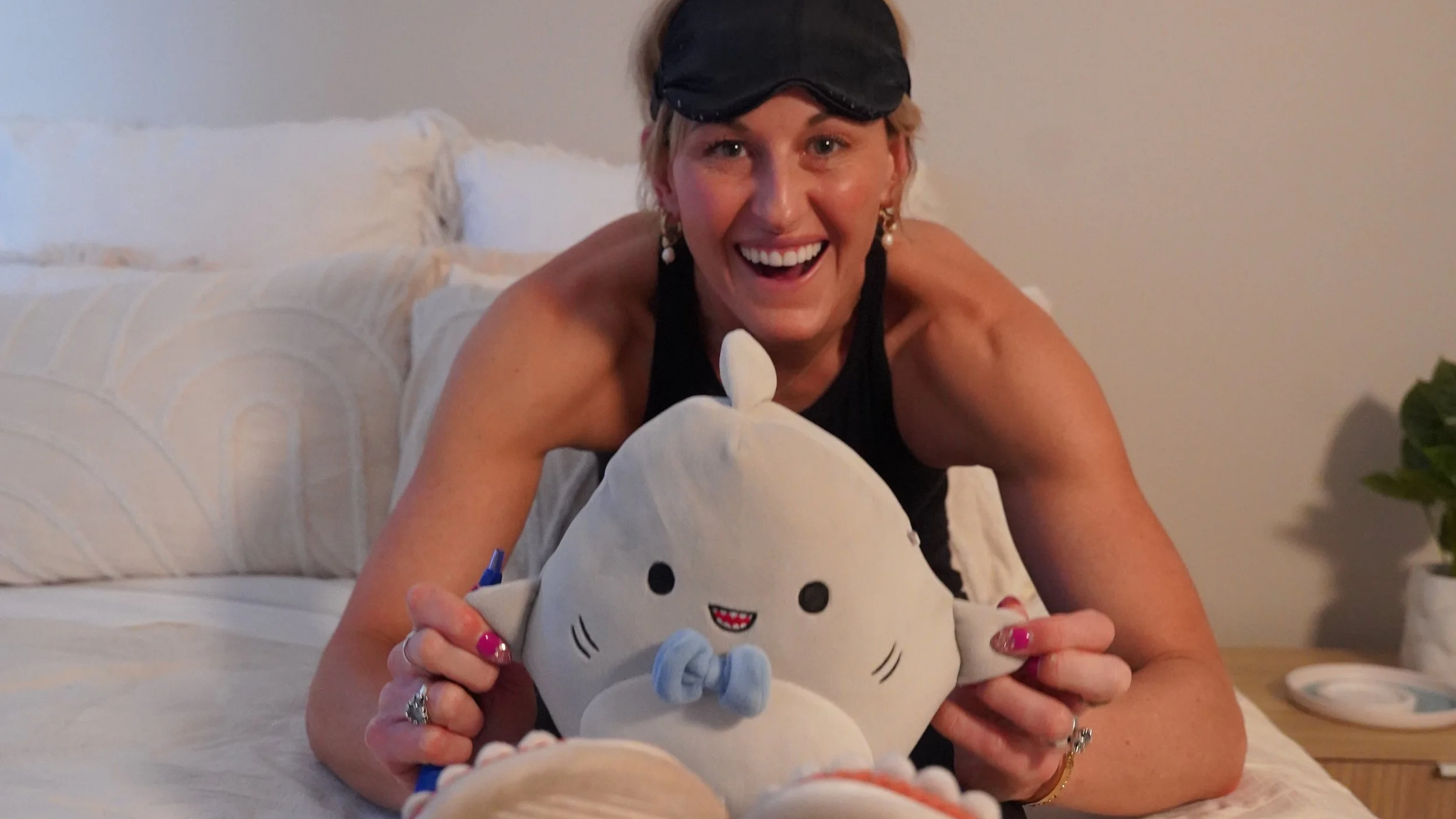 A woman with short blonde hair, wearing a black sleep mask on her forehead, smiling broadly. She is sitting on a bed with white and beige bedding, holding a plush toy of a white cat with a blue bow and a smiling face. She has multiple rings on her fingers and earrings, and is wearing a black sleeveless top.