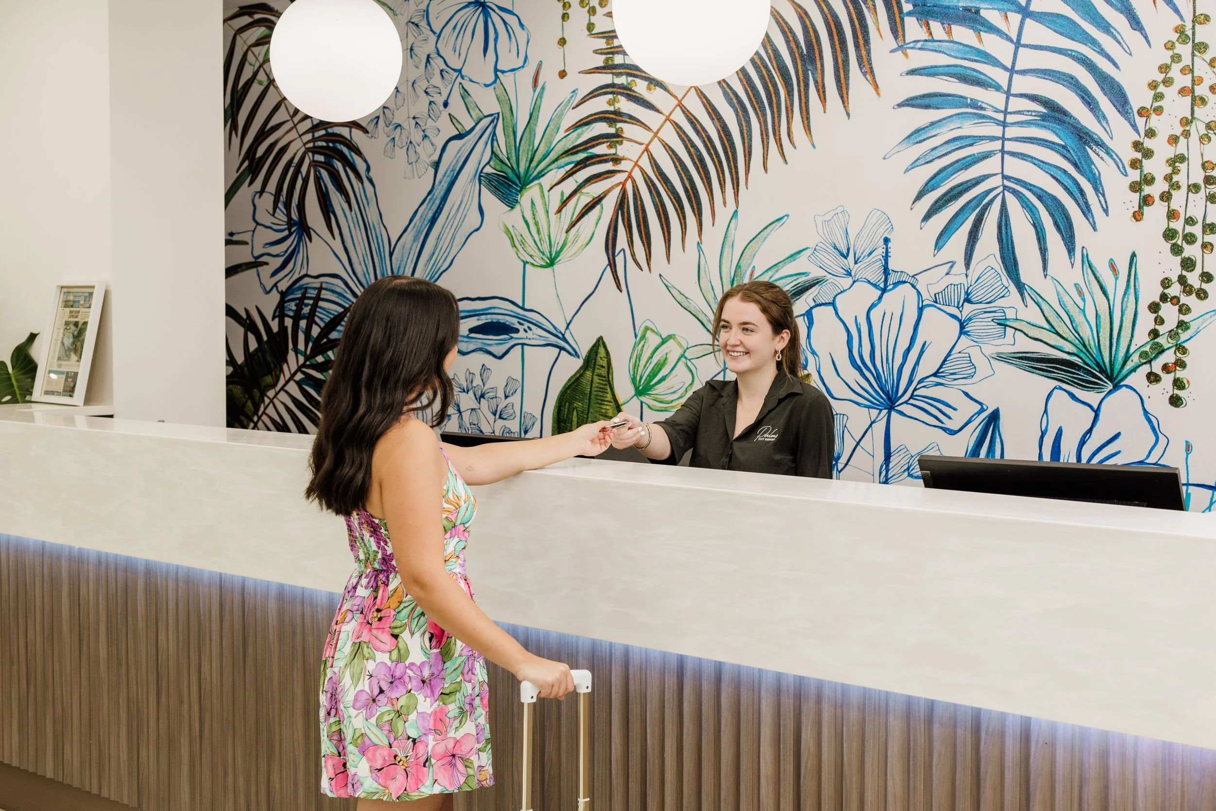 A woman in a floral dress check-in at a hotel front desk with a smiling receptionist against a decorative blue, green, and black tropical leaf mural.