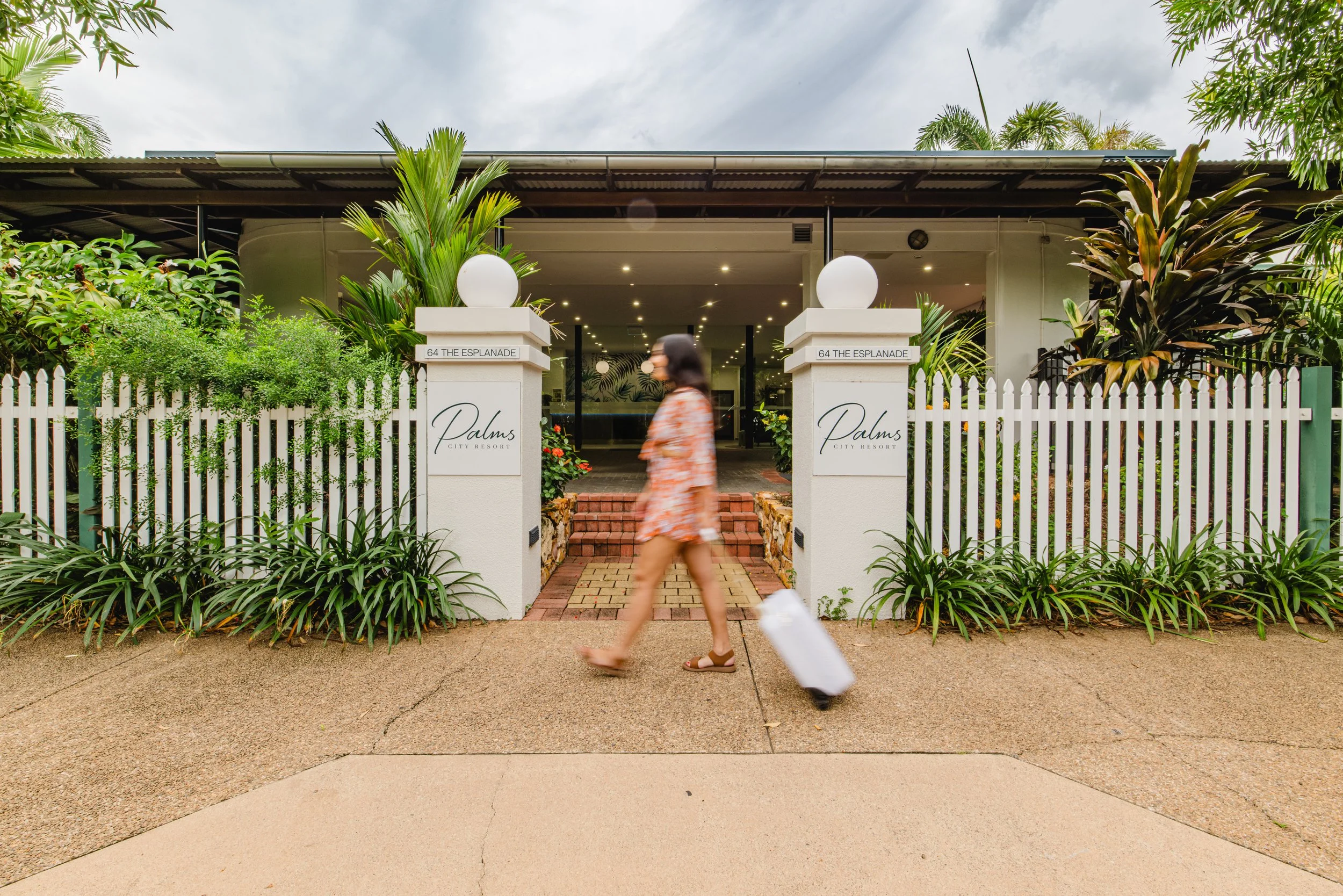 A woman walking with a rolling suitcase past the entrance of Palms City Resort, which is surrounded by greenery and white fencing.