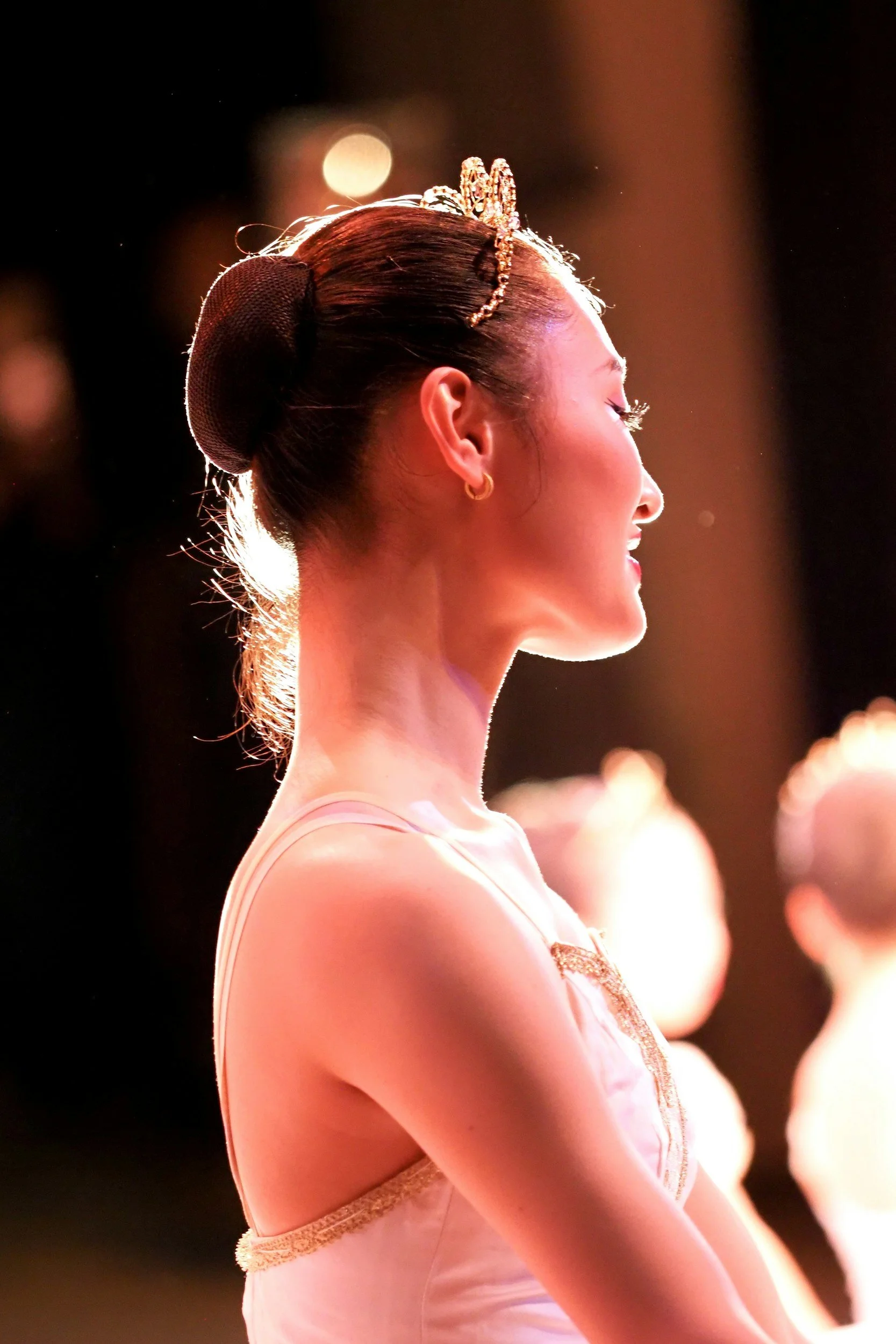 Close-up side profile of a ballerina with her eyes closed, wearing a tiara and a ballet costume with gold accents, illuminated by stage lighting about to compete in the AABC Competition