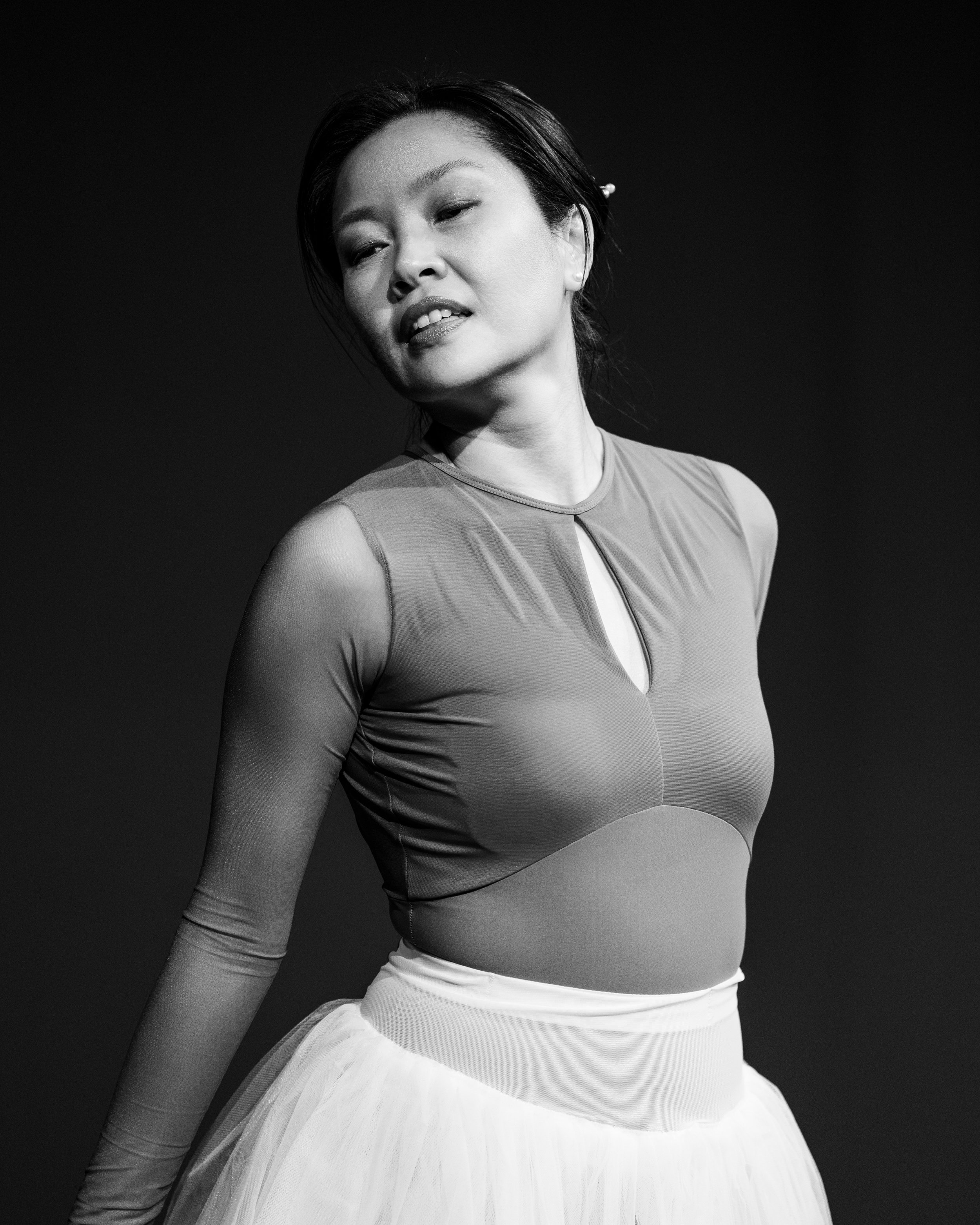 Black and white photograph of an Adult Ballet Centre dancer in a sleeveless top and a light-colored skirt, with her head tilted slightly to the side and her eyes partially closed in her performance