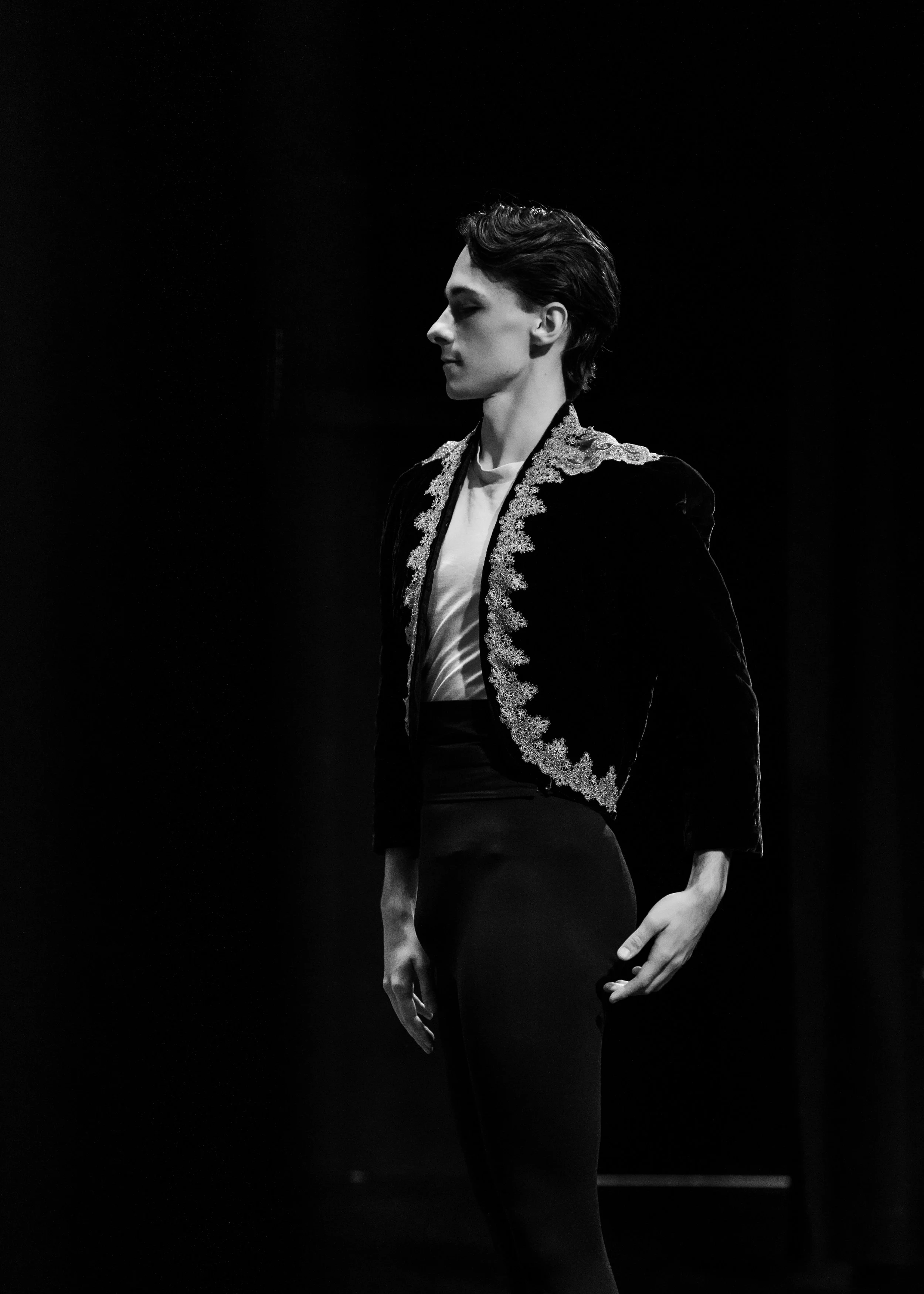 Black and white photo of a dancer with short hair wearing a prince jacket with ornate embroidery, standing sideways with eyes closed, against a dark background, performance ready for the AABC Competition