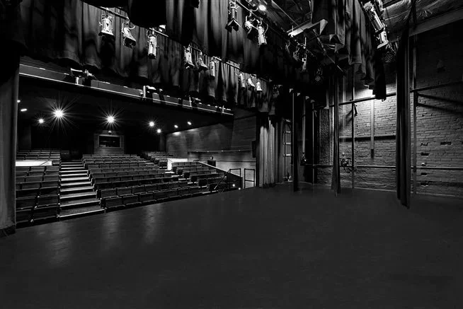 Shirley Burke Theatre empty, a balck and white photo from side stage curtains, hanging stage lights, and an empty auditorium with rows of seats, before the AABC Competition begins.